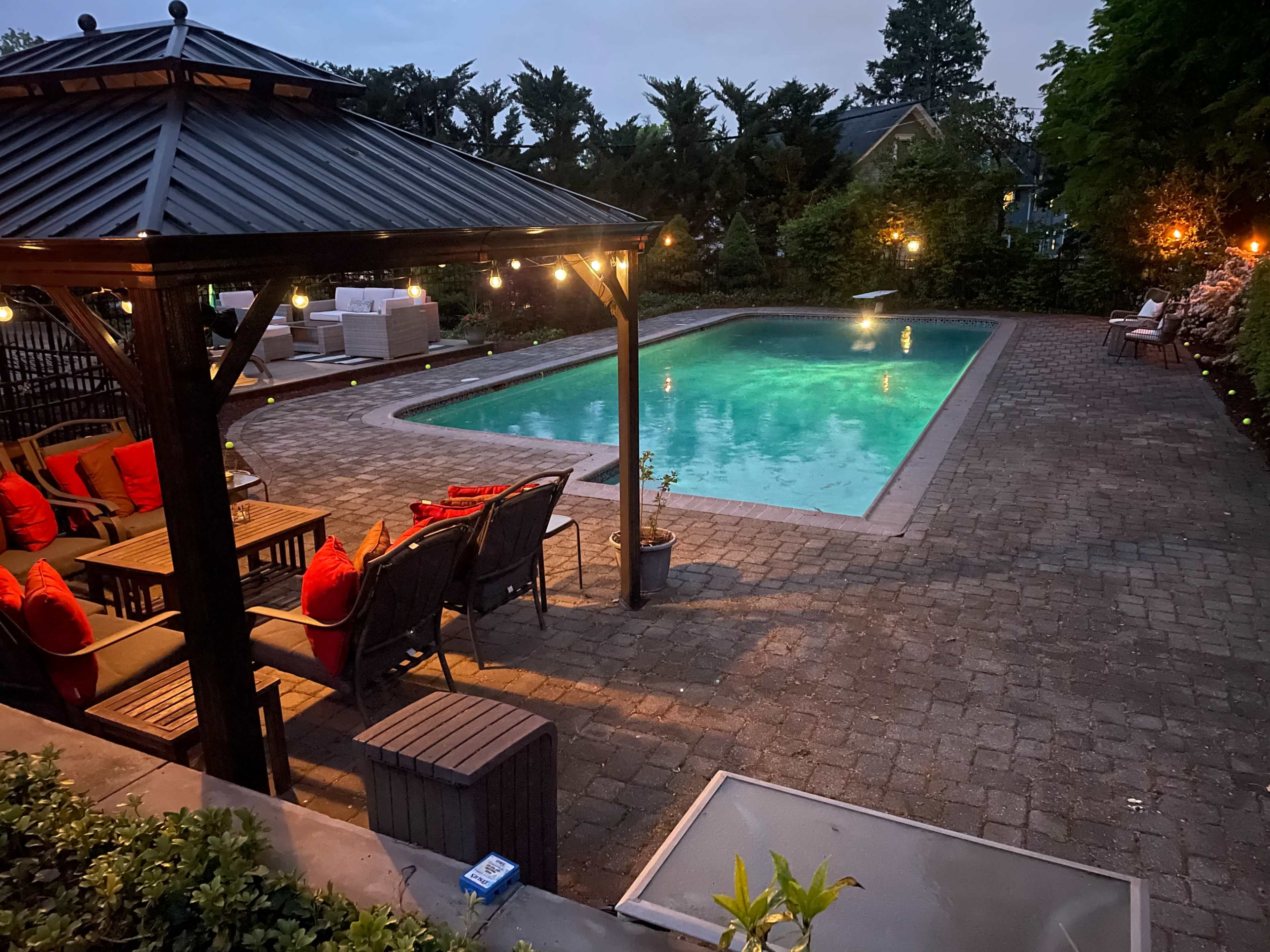 The image shows a backyard pool area featuring a rectangular swimming pool, surrounded by paved stones and a gazebo with seating and decorative lighting.