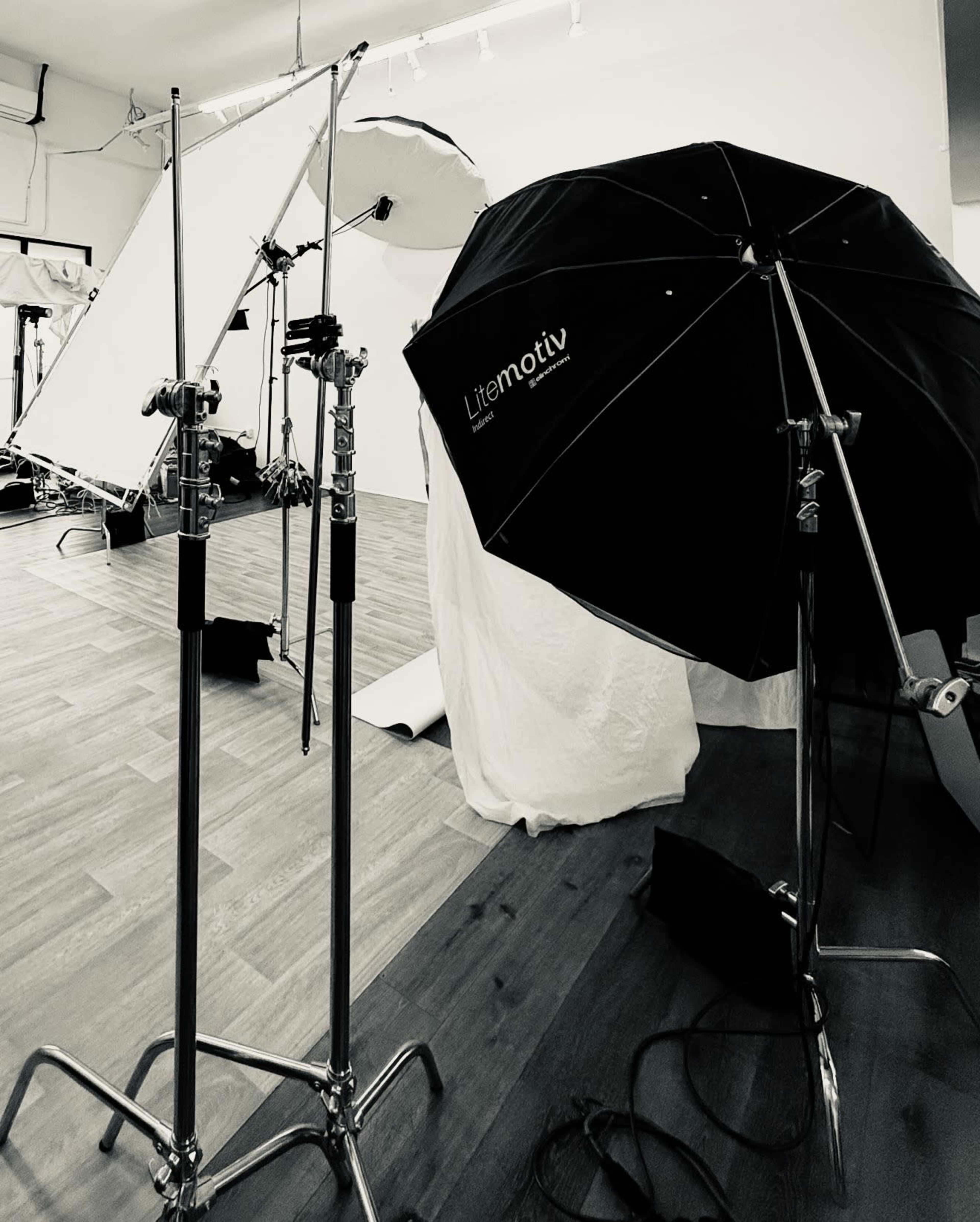 The image shows several photography equipment stands, including a black umbrella reflector, set up in a studio space with wooden flooring and white backdrop fabric.