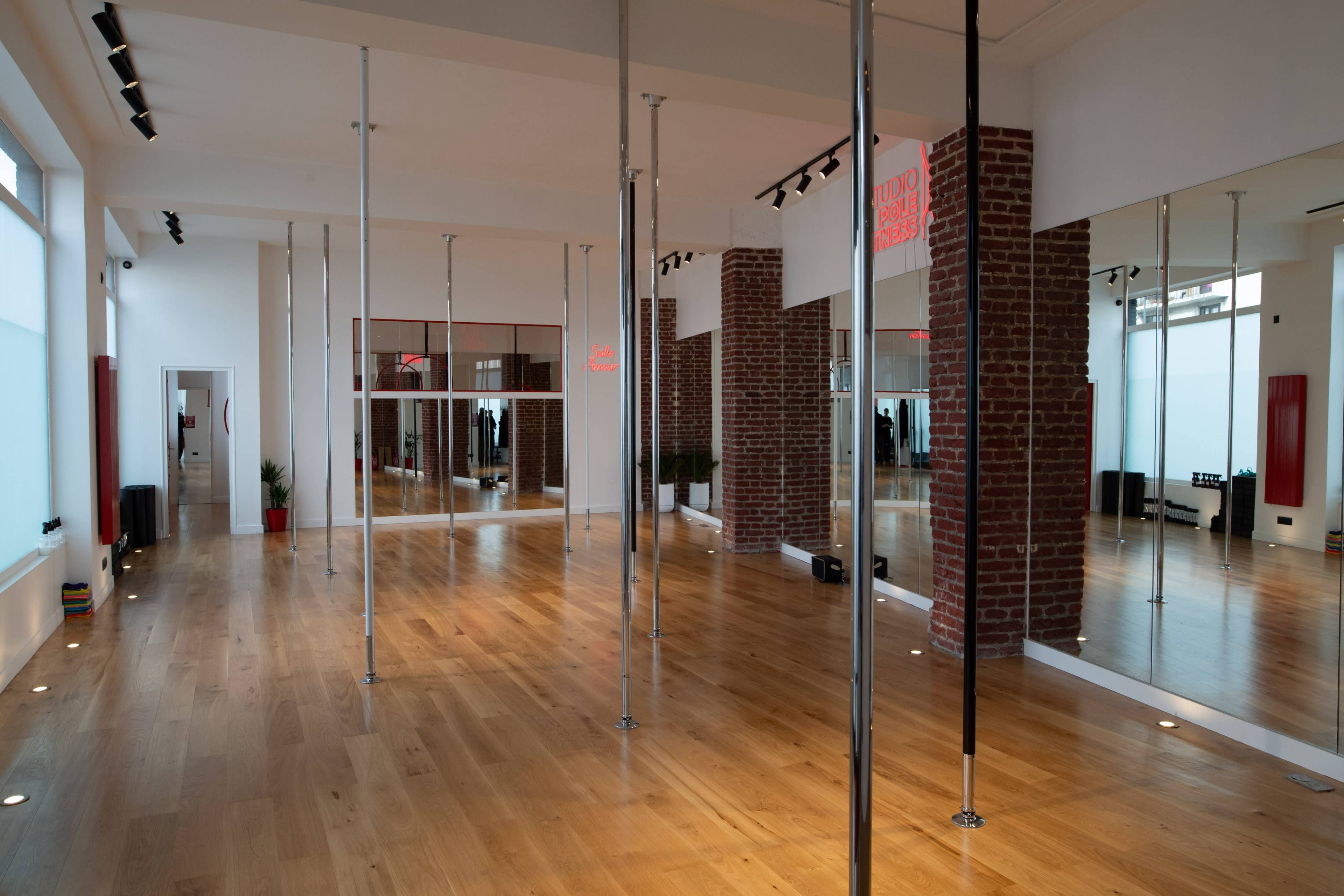The image shows a spacious dance studio with wooden flooring, mirrored walls, and several vertical poles.