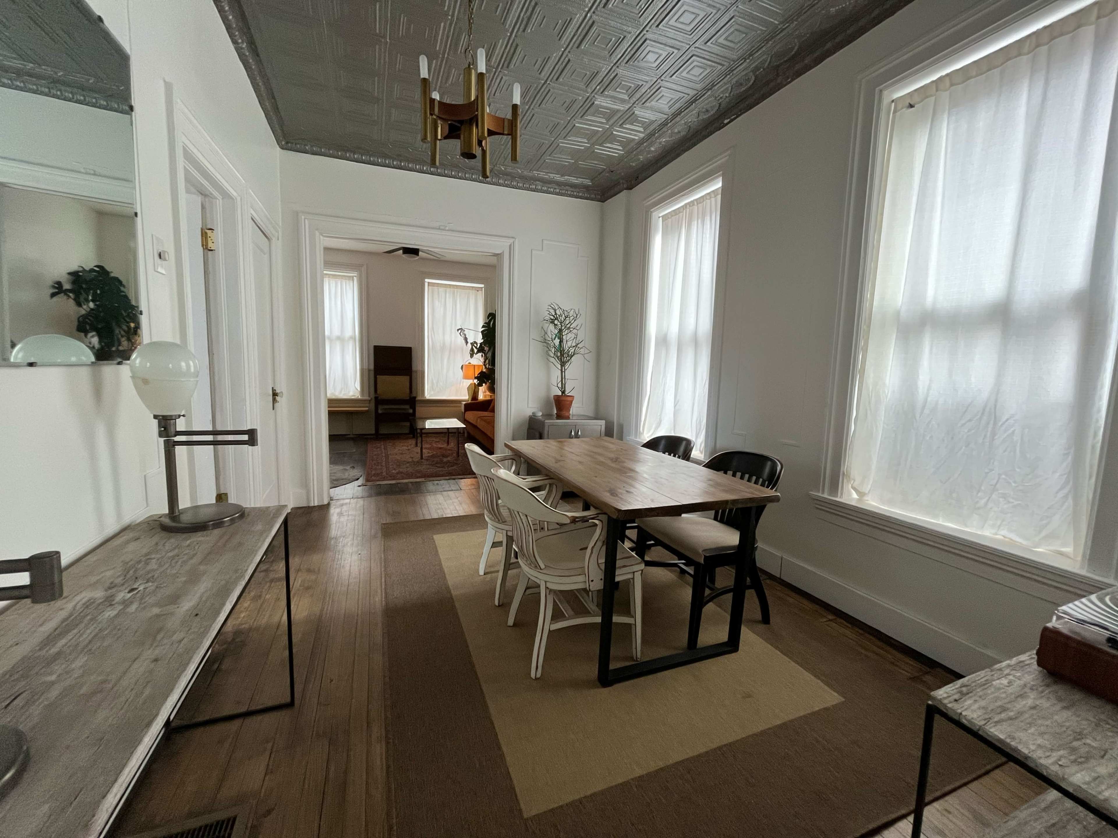 A dining area with a long wooden table surrounded by chairs, a side console table, and large windows with sheer curtains.