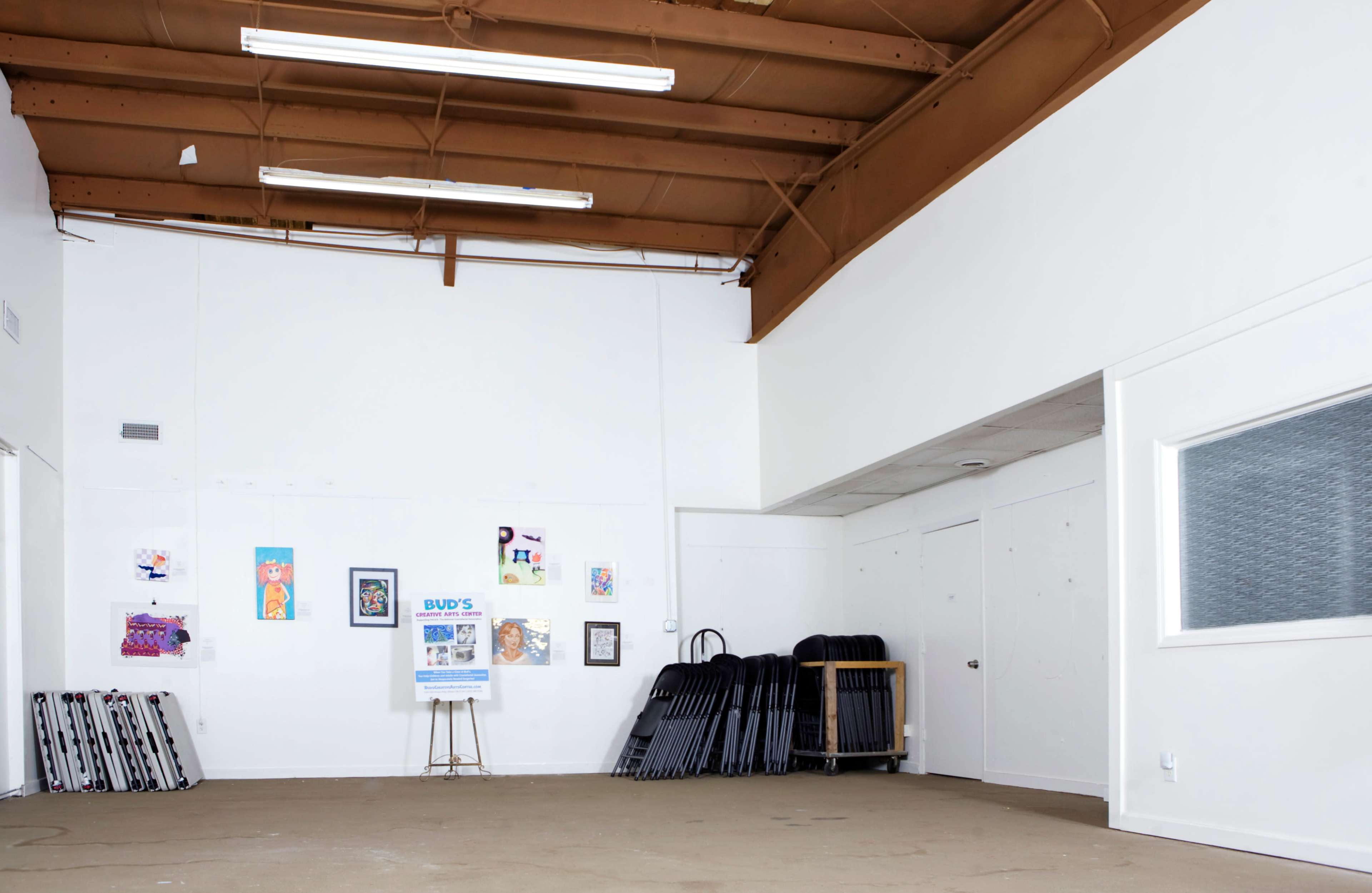 The image shows an empty room with a high ceiling, featuring artwork on the wall, a small easel, and several stacked chairs against one side.