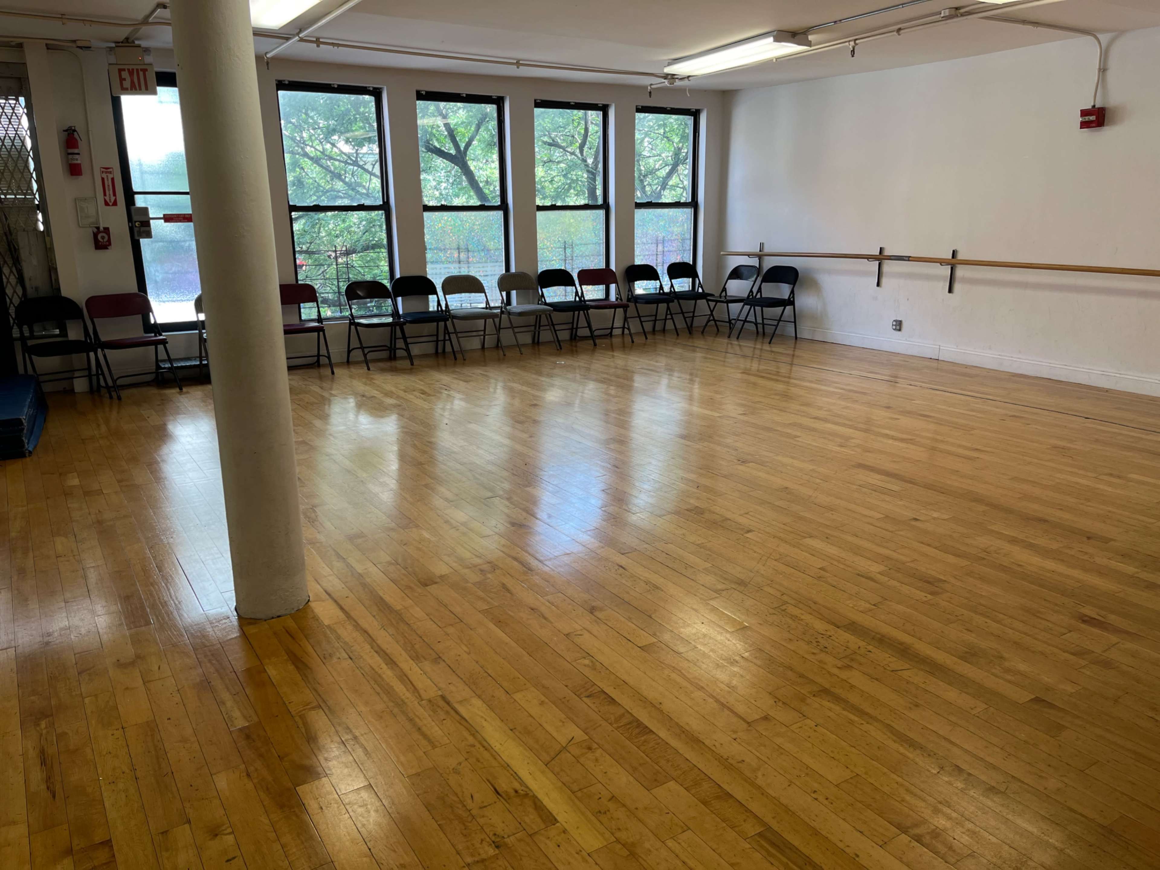 A dance studio with a polished wooden floor, a row of chairs against the wall, and large windows letting in natural light.
