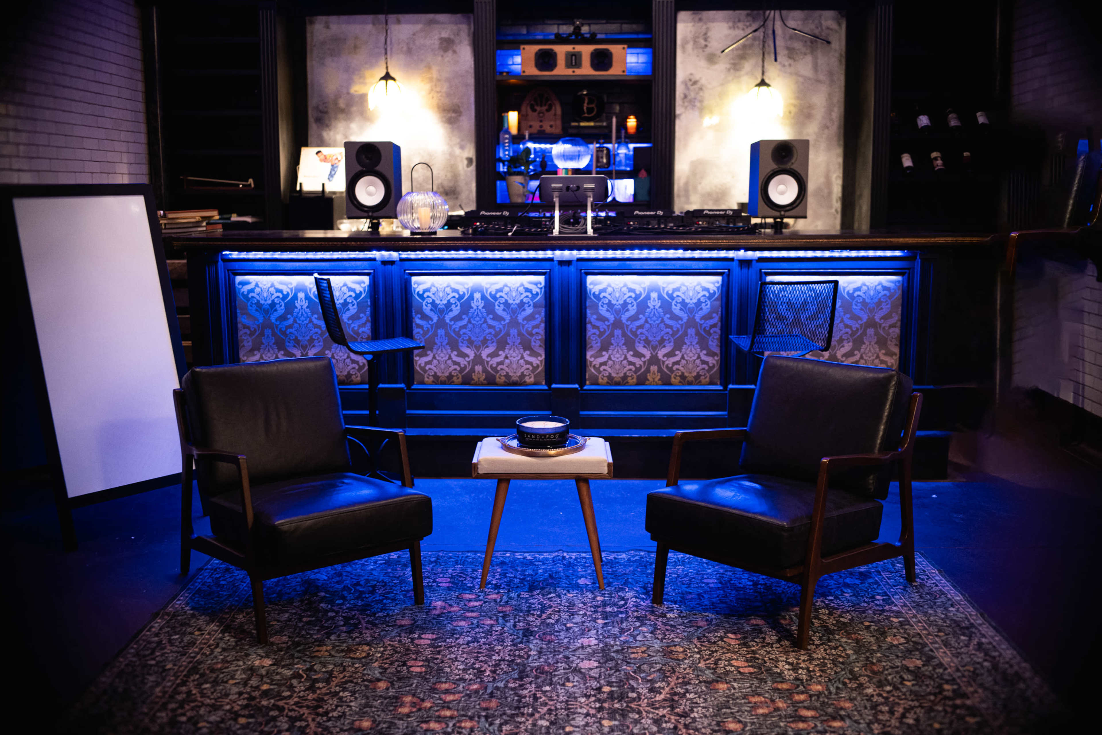 The image shows a dimly lit lounge area featuring two black chairs facing a small table, with a bar and audio setup illuminated by blue lighting in the background.