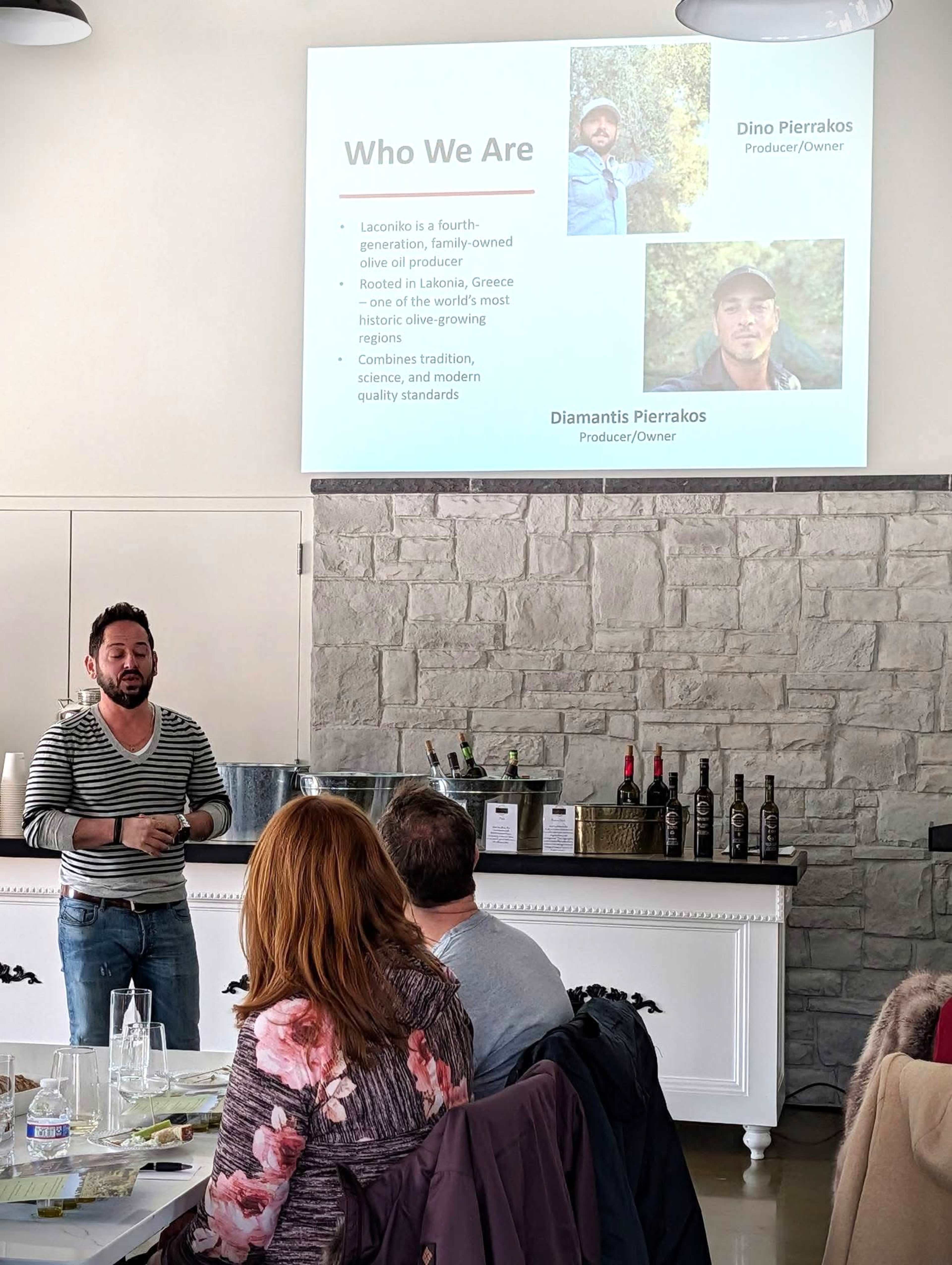 A presentation is taking place in a bright room, featuring a speaker discussing a family-owned olive oil business, while an audience looks on.