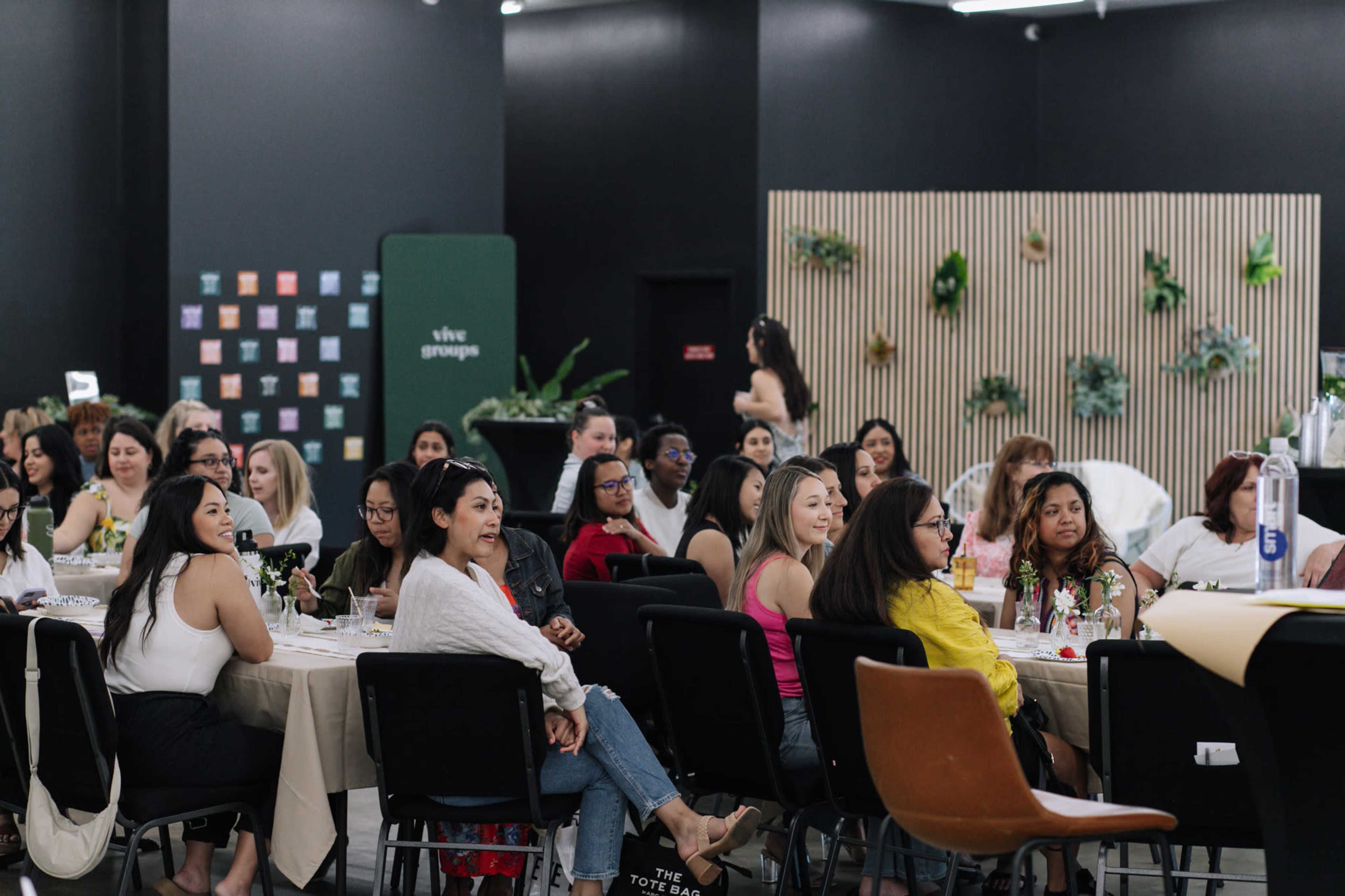 A diverse group of people is seated at tables in a well-decorated event space, engaged in conversation.