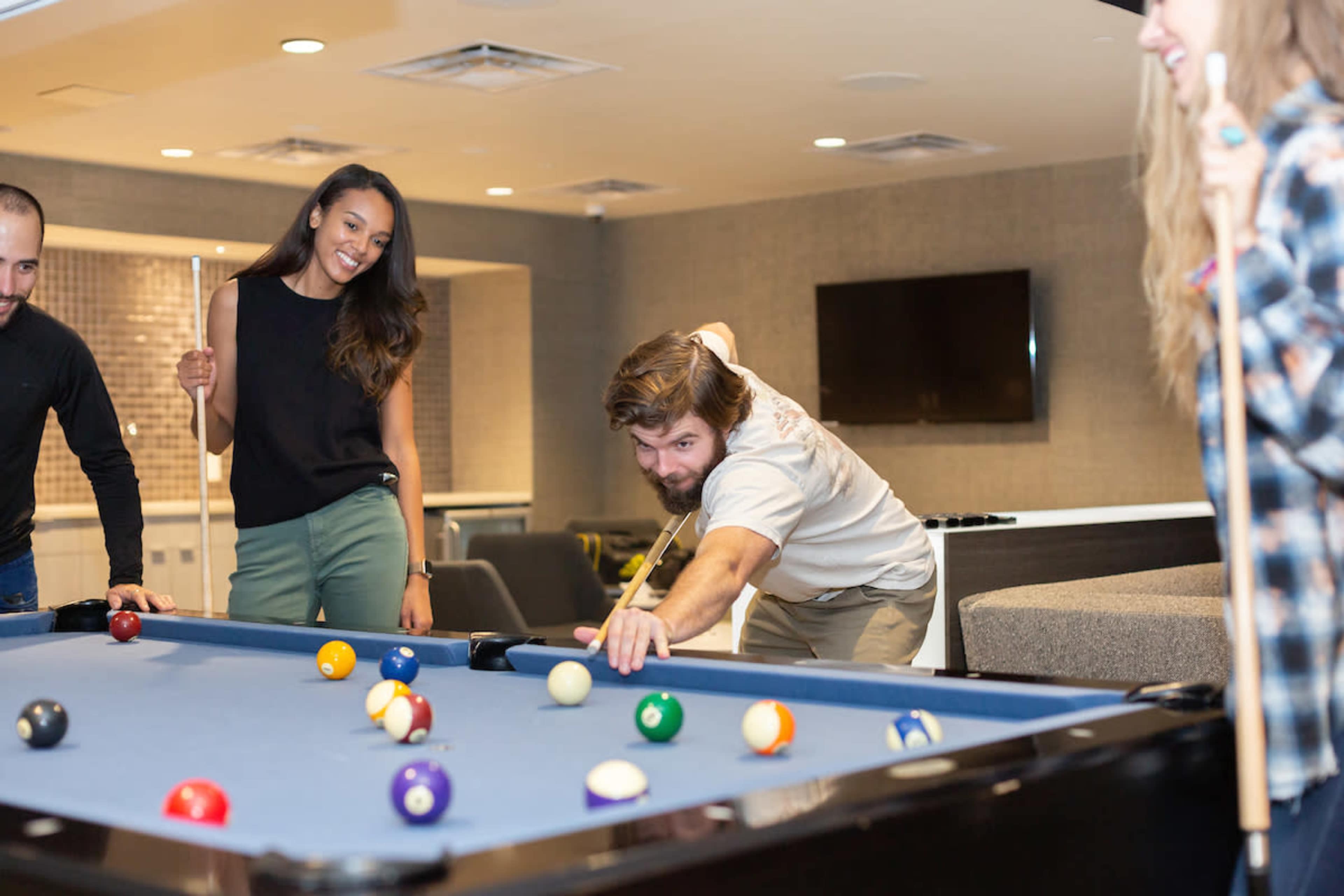 A group of four people play pool in a recreational room with a blue billiard table.