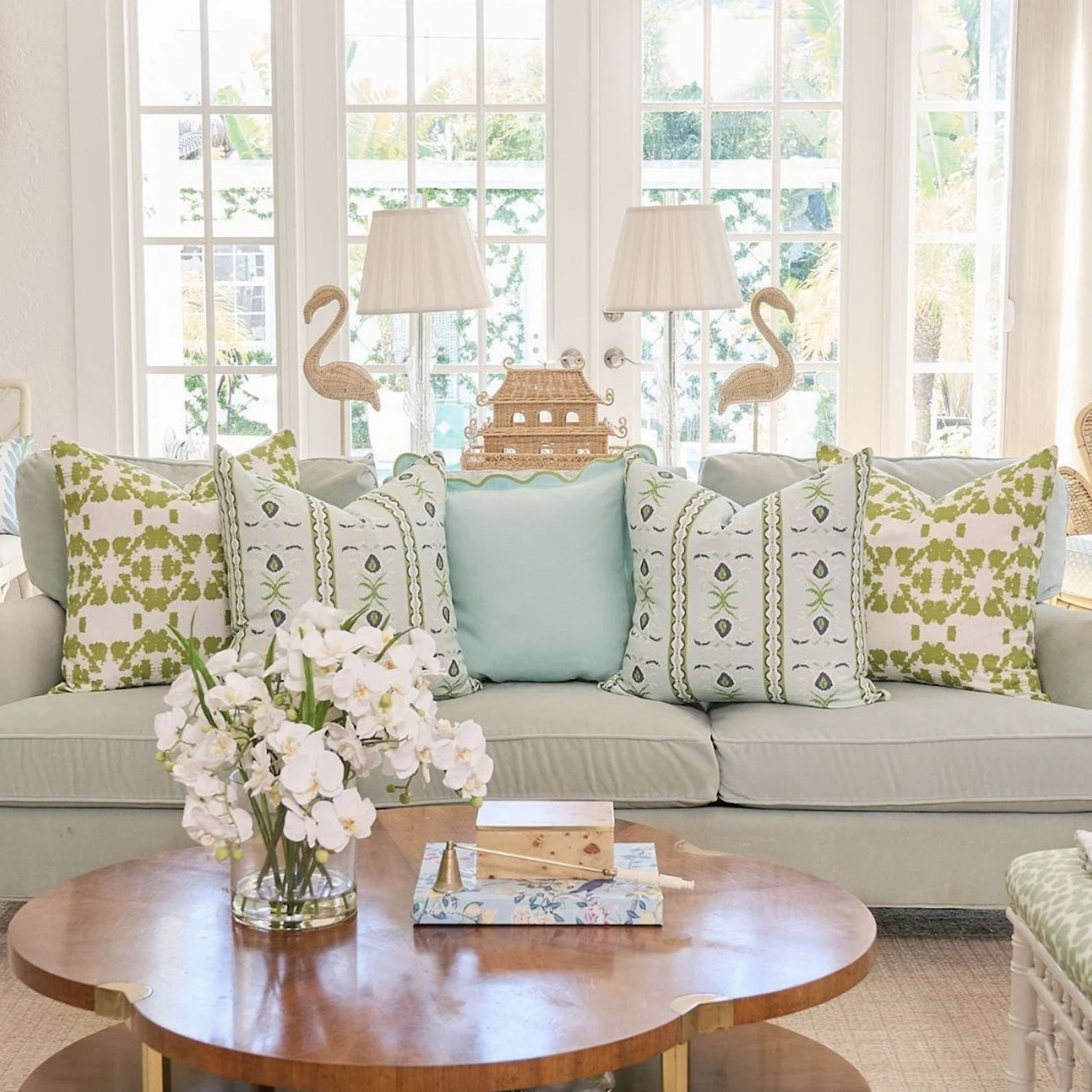 A light green sofa with patterned pillows is positioned in a bright room filled with natural light, alongside a wooden table and decorative accents.