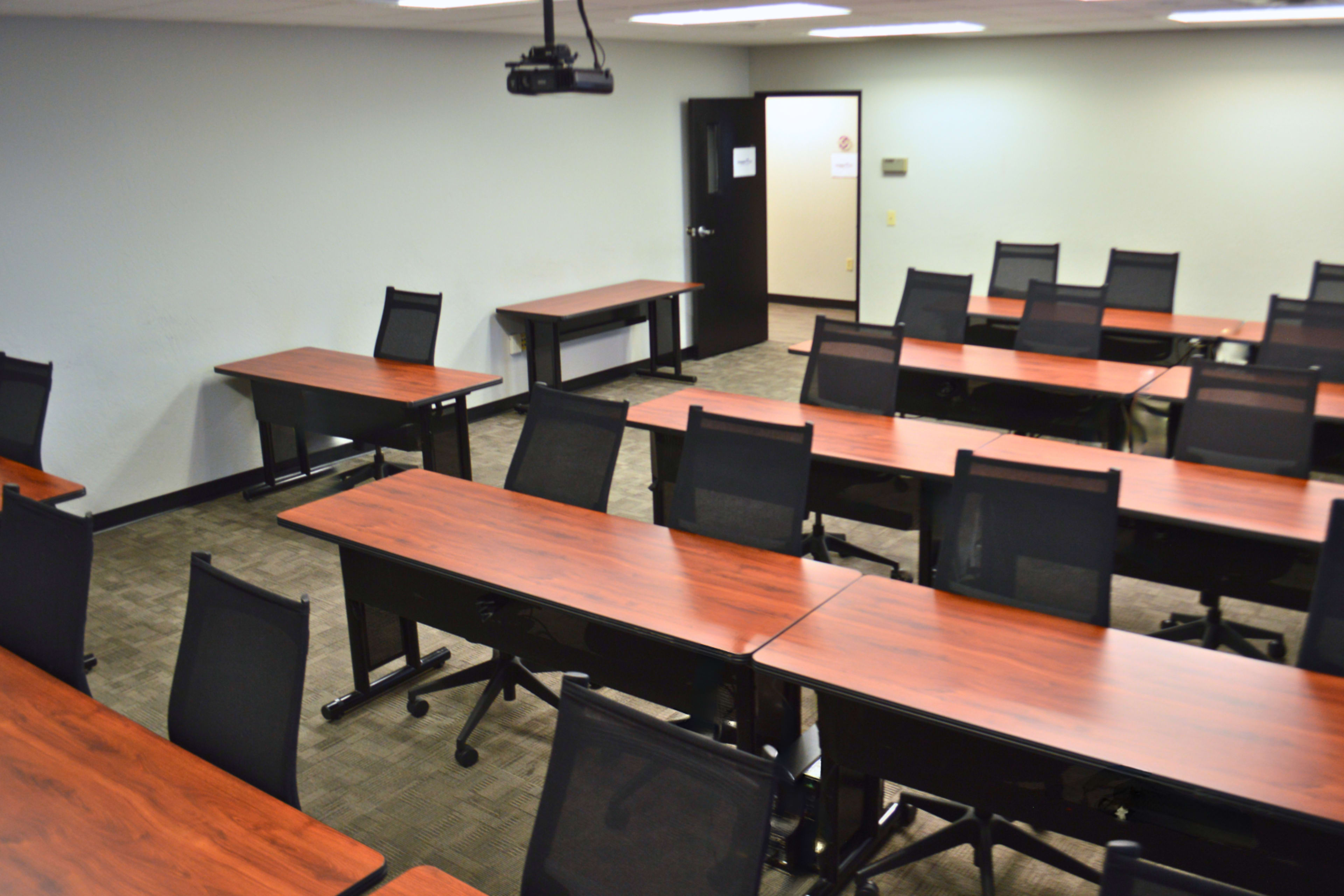 A classroom is set up with multiple rows of wooden tables and black mesh chairs arranged for a lecture or meeting.