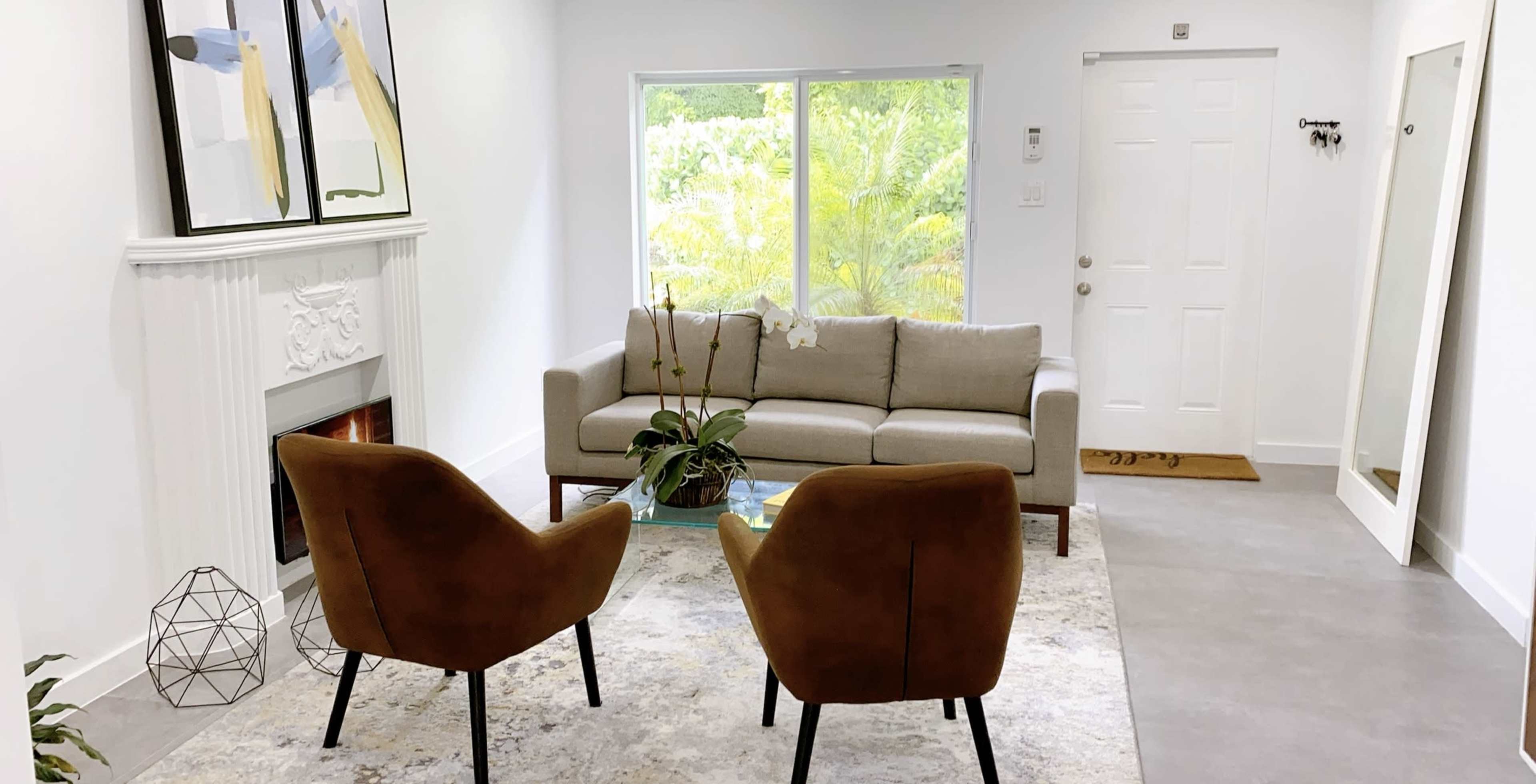 The image shows a modern living room featuring a gray sofa, two brown chairs, a decorative plant, and a large window with greenery outside.