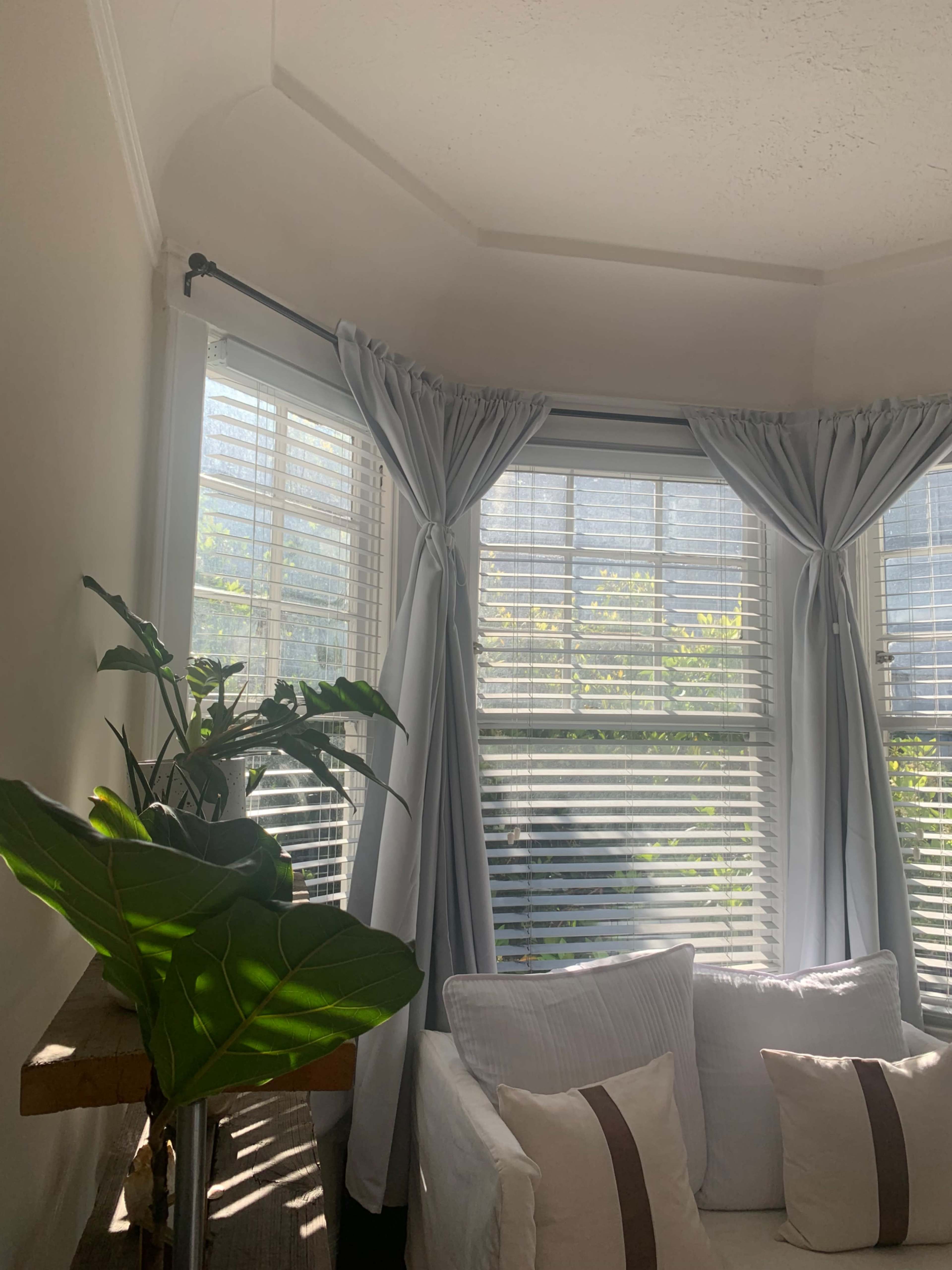 A bright corner of a room features large windows with sheer curtains, allowing sunlight to illuminate a comfortable white sofa and a potted plant.
