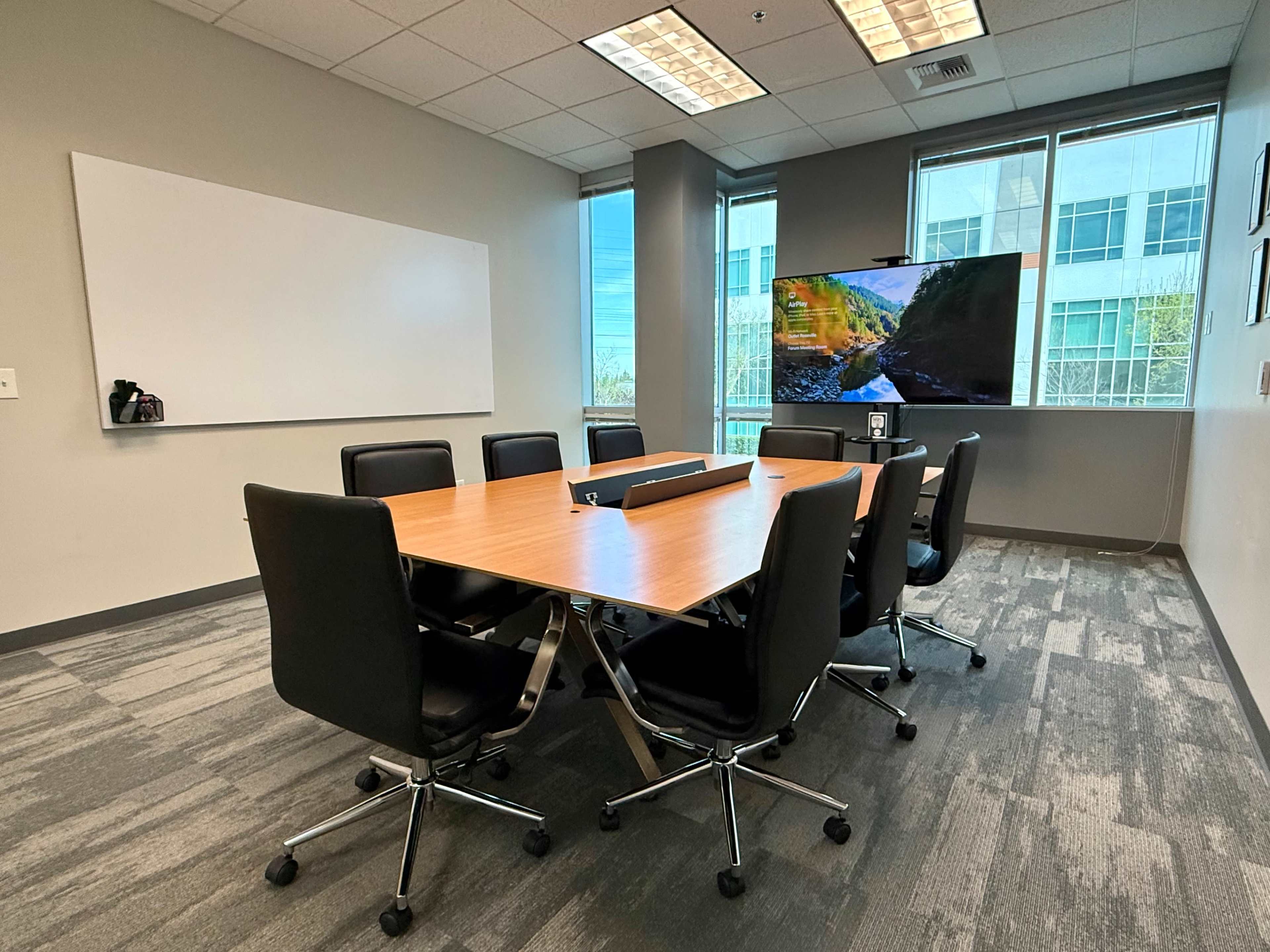 The image shows a modern conference room with a large table surrounded by black chairs, a whiteboard, and a wall-mounted screen displaying a scenic landscape.