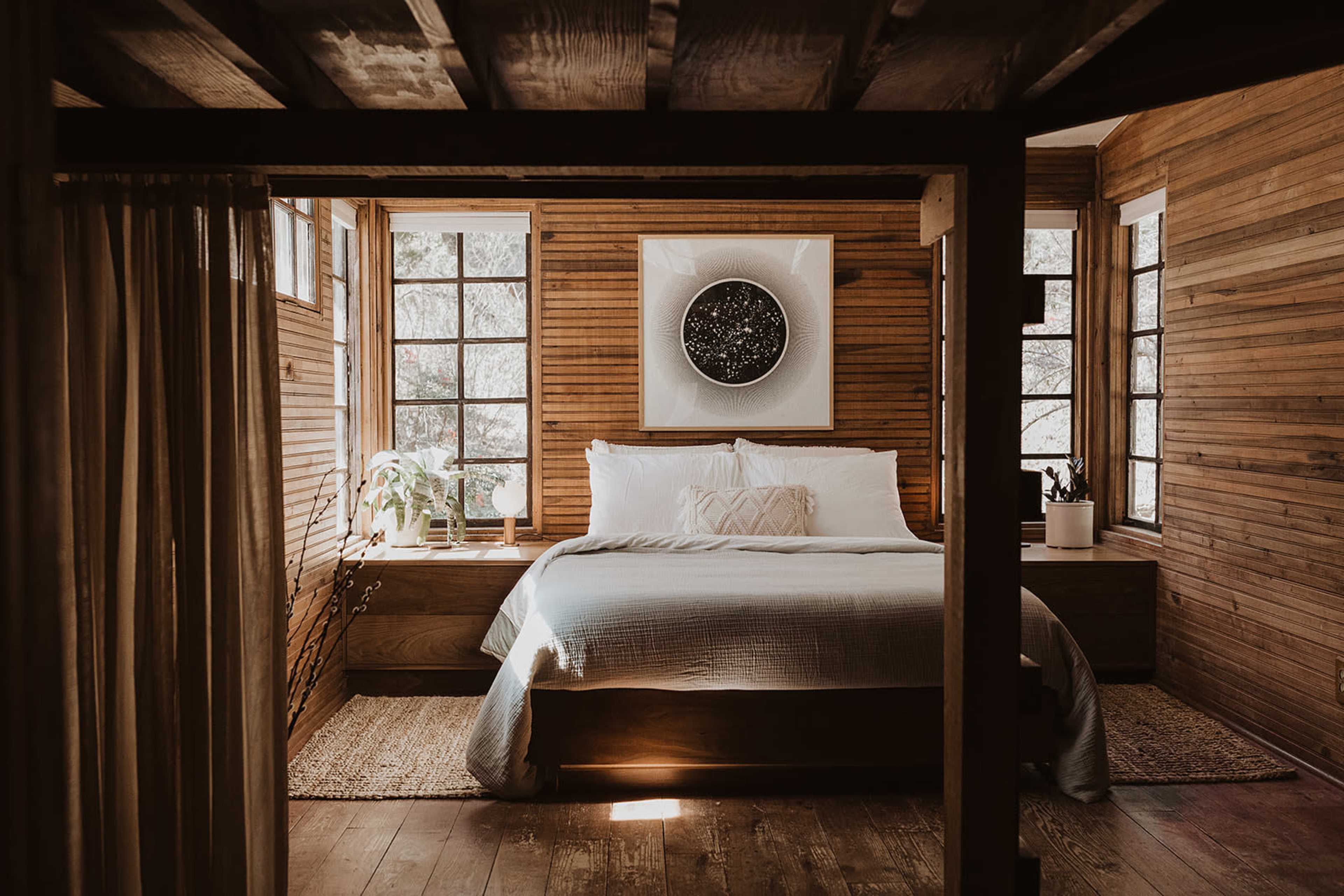 A cozy bedroom with wooden walls, a large bed in the center, and large windows allowing natural light to enter.