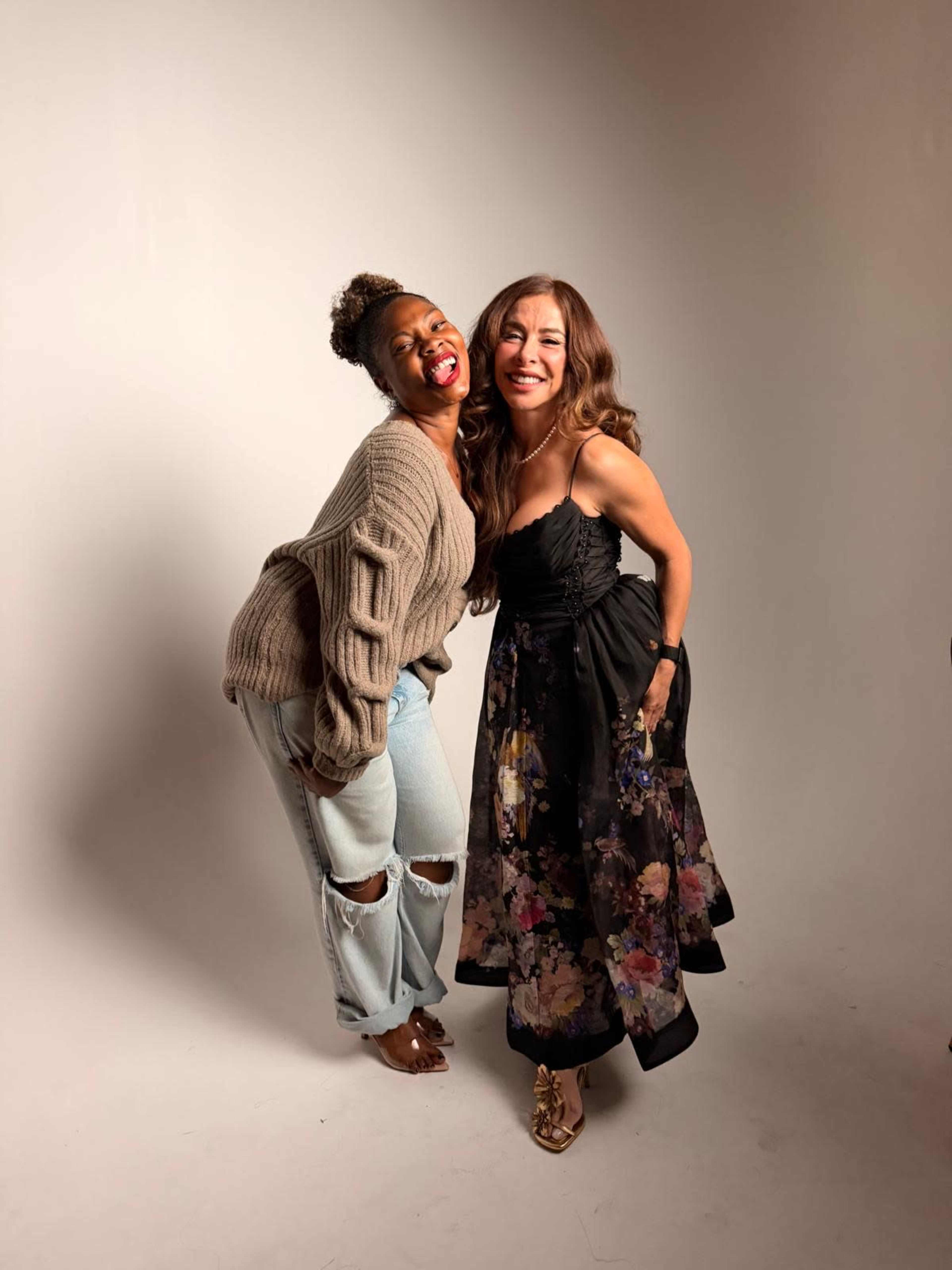 Two women pose together, one in a cozy sweater and jeans and the other in a floral-printed dress, against a neutral background.