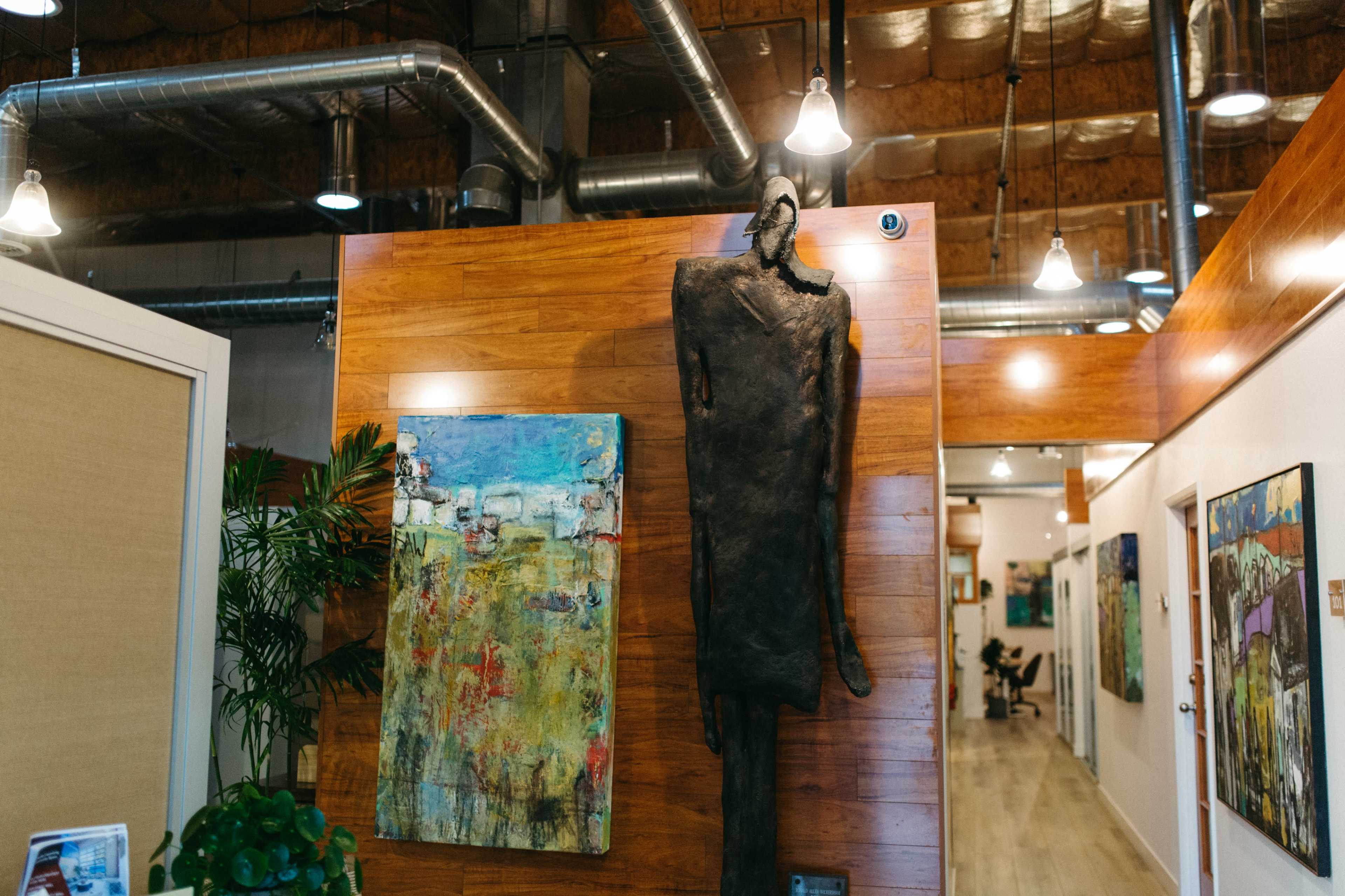 The image shows a modern interior space with wooden walls, featuring a large abstract painting and a sculpture mounted on the wall.