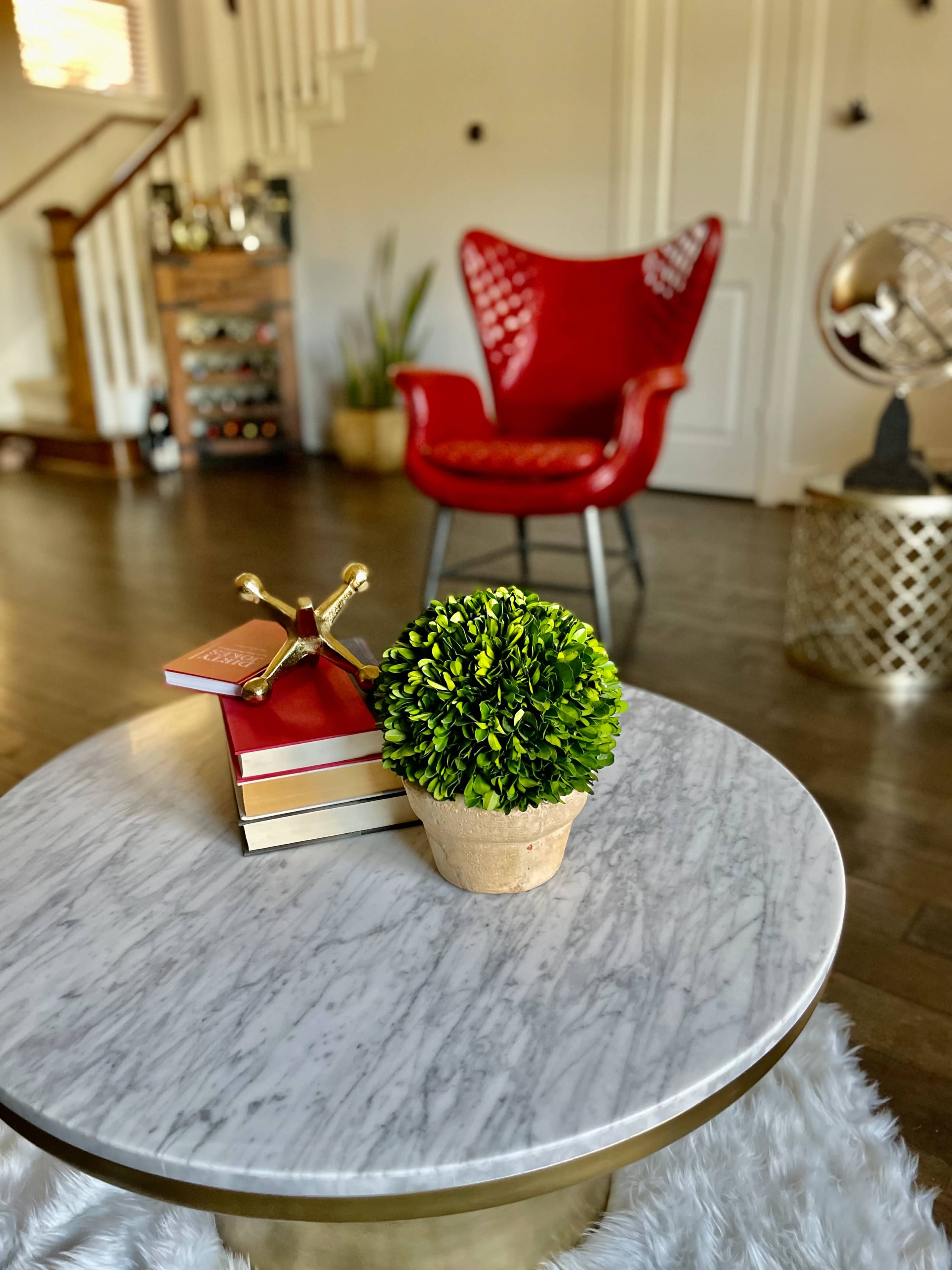 A marble-top coffee table holds stacked books and a small potted plant beside a red chair in a well-decorated living space.