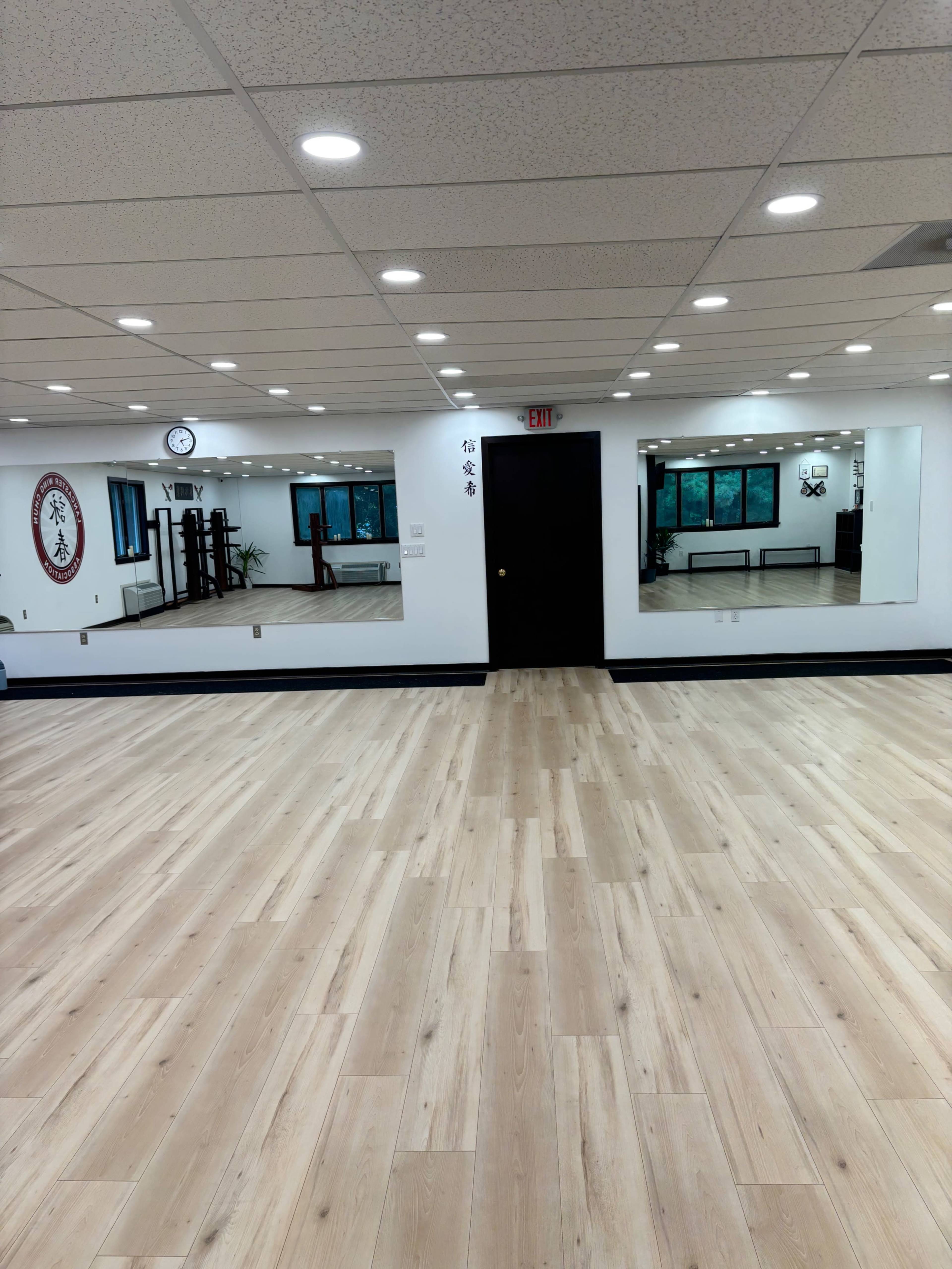 The image shows a well-lit martial arts studio with wooden flooring, two mirrored walls, and a clock on the wall.