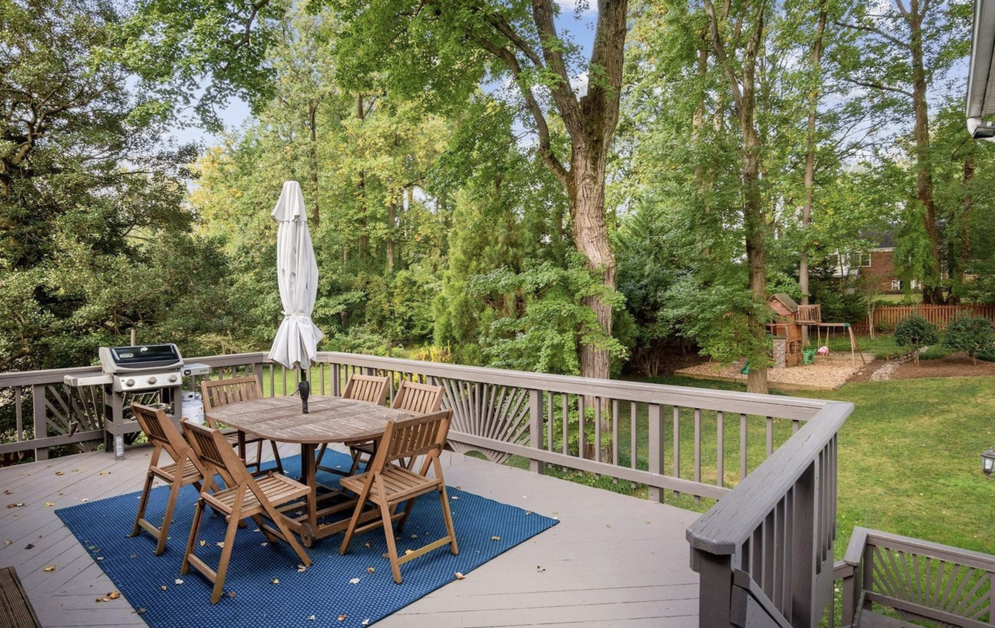 A wooden deck features a circular table with six chairs, an umbrella, and a grill, overlooking a lush green backyard with trees and play structures.