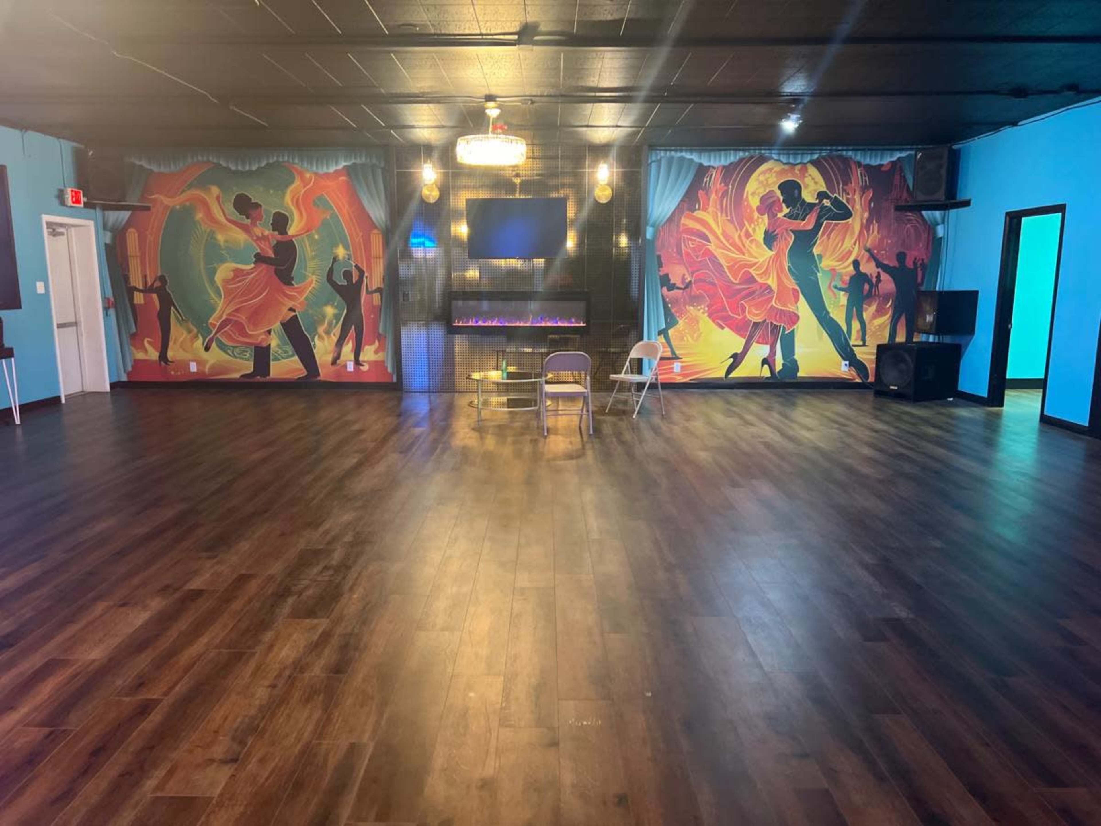 The image shows a spacious dance studio with colorful murals on the walls, a large wooden floor, and a modern electric fireplace.