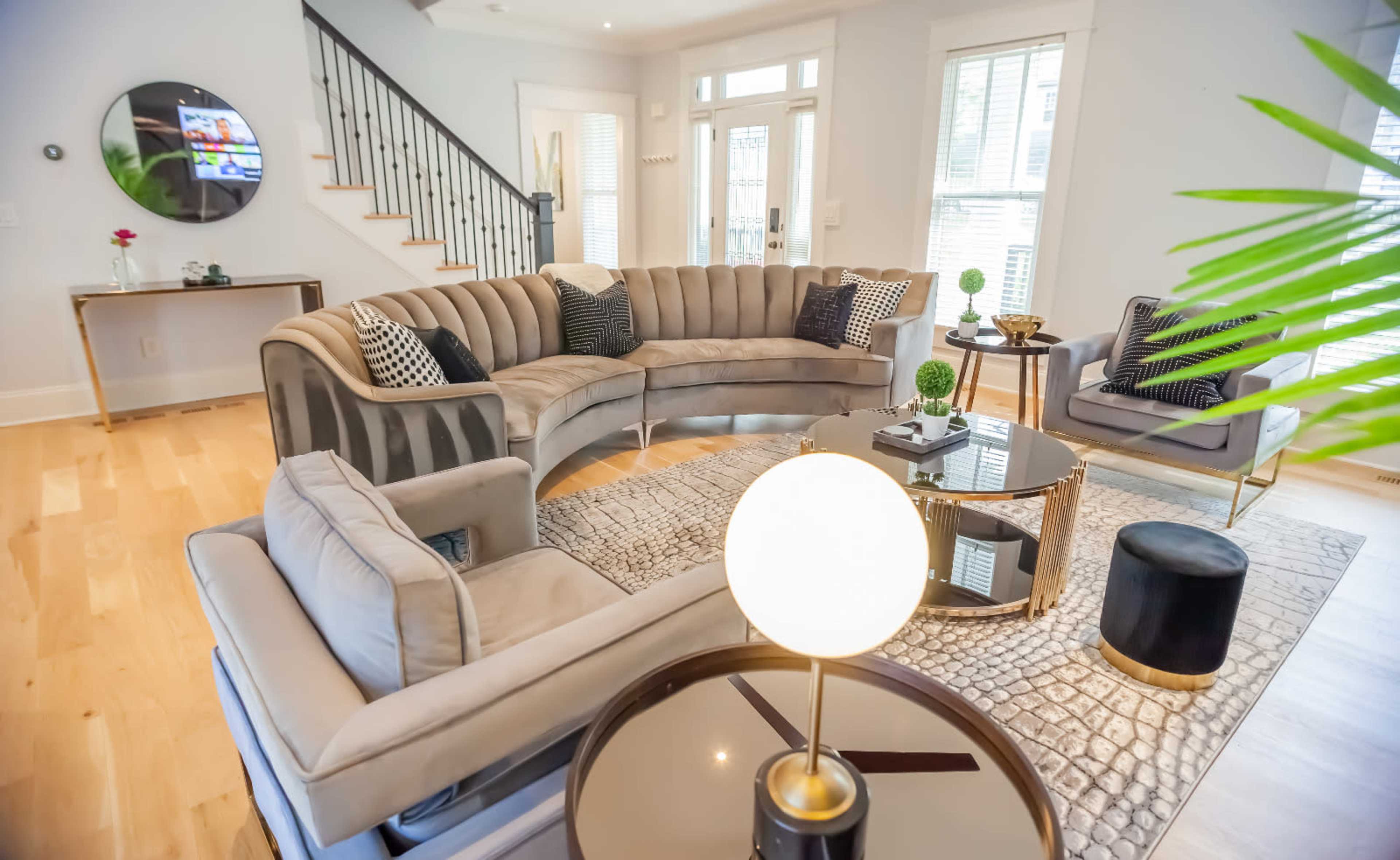 A spacious living room features a large curved sofa, a round coffee table, and two accent chairs, complemented by natural light from multiple windows.