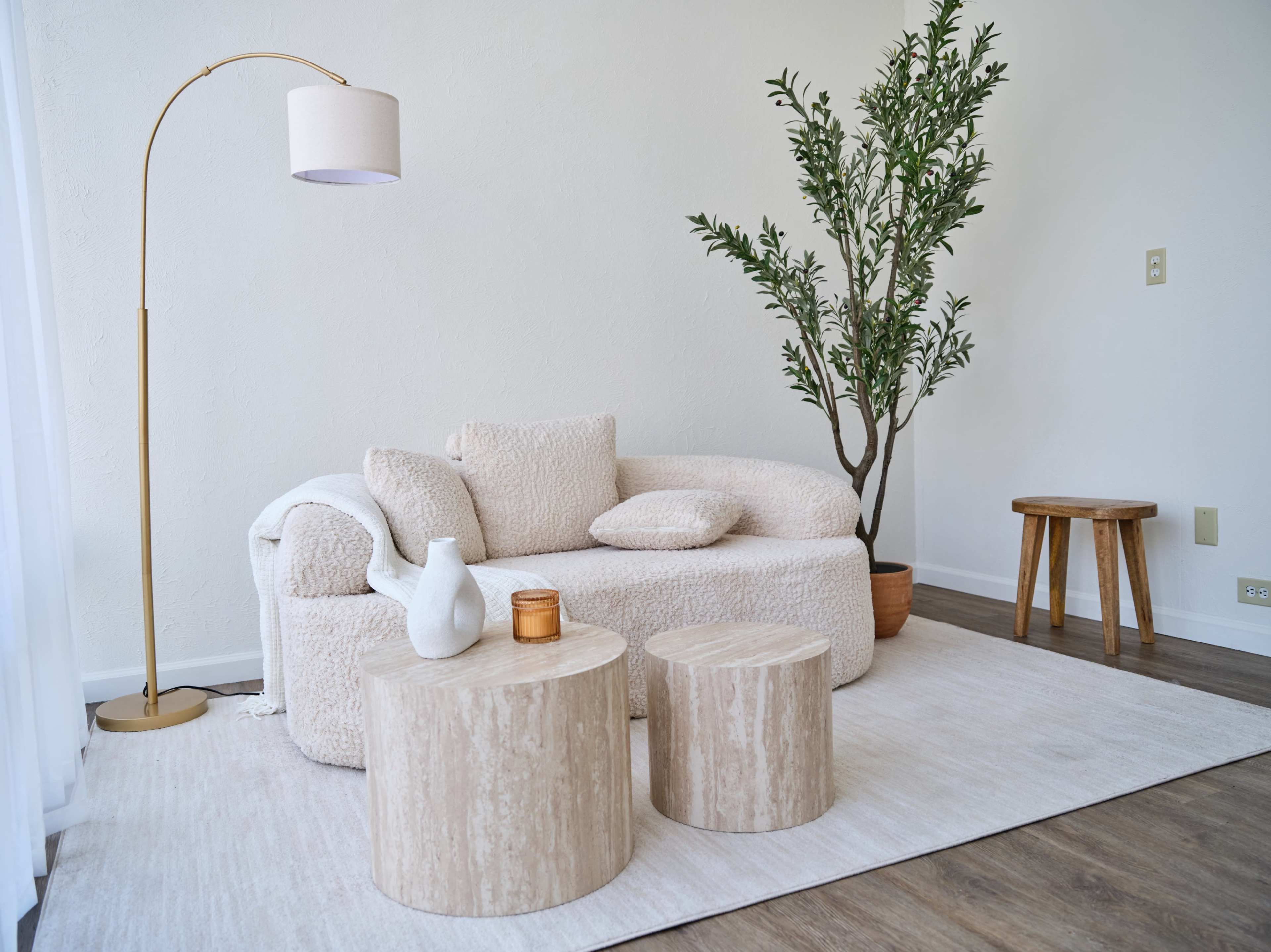 The scene features a cozy living room corner with a soft, beige sofa, two round wooden coffee tables, and a tall floor lamp, accompanied by a potted plant against a white wall.
