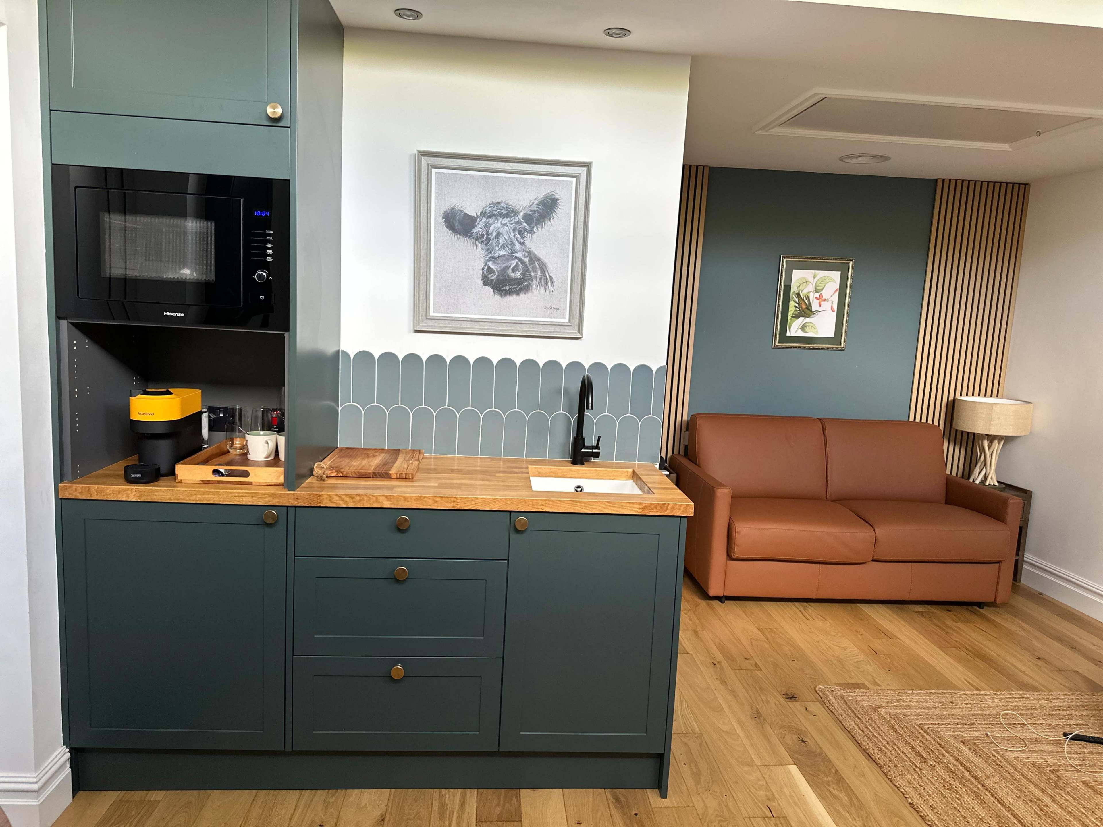 The image shows a modern kitchen area with teal cabinetry, a wooden countertop, a microwave, and a living space featuring a brown sofa and decorative wall art.