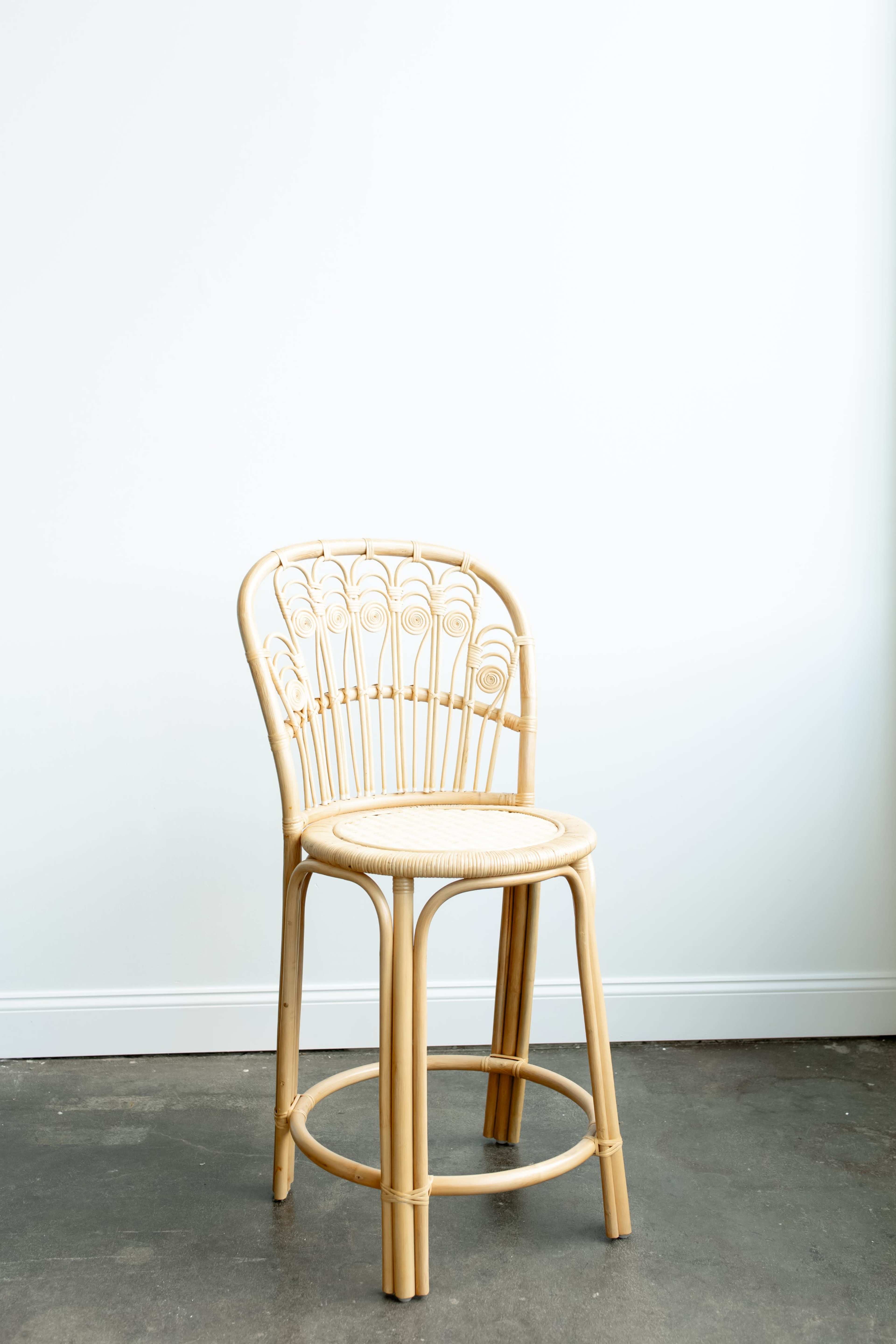 A woven rattan bar stool with a circular seat and a curved backrest stands against a plain white wall.