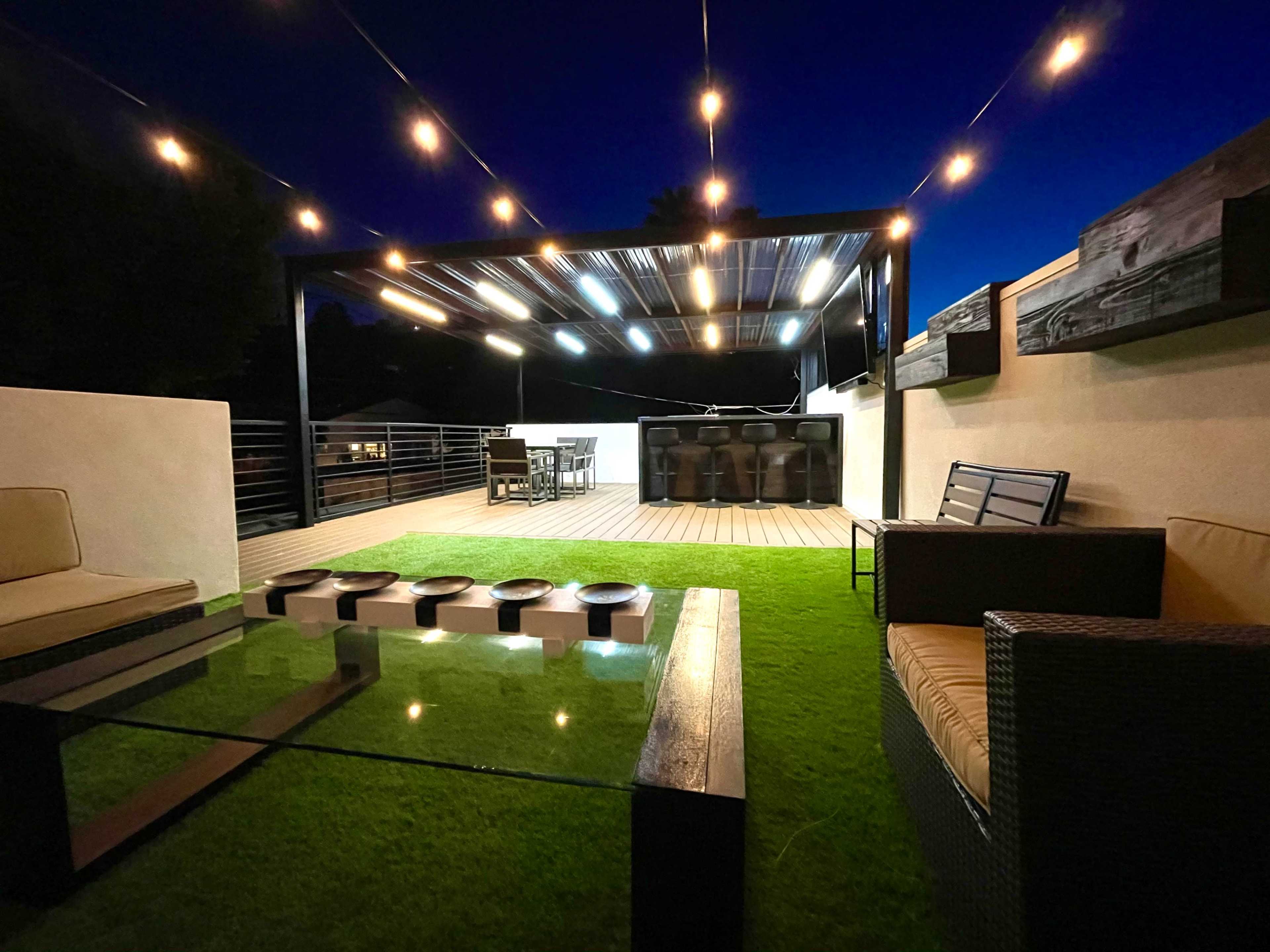 A rooftop terrace features a seating area with a glass coffee table, surrounded by green turf and illuminated by strung lights overhead at night.
