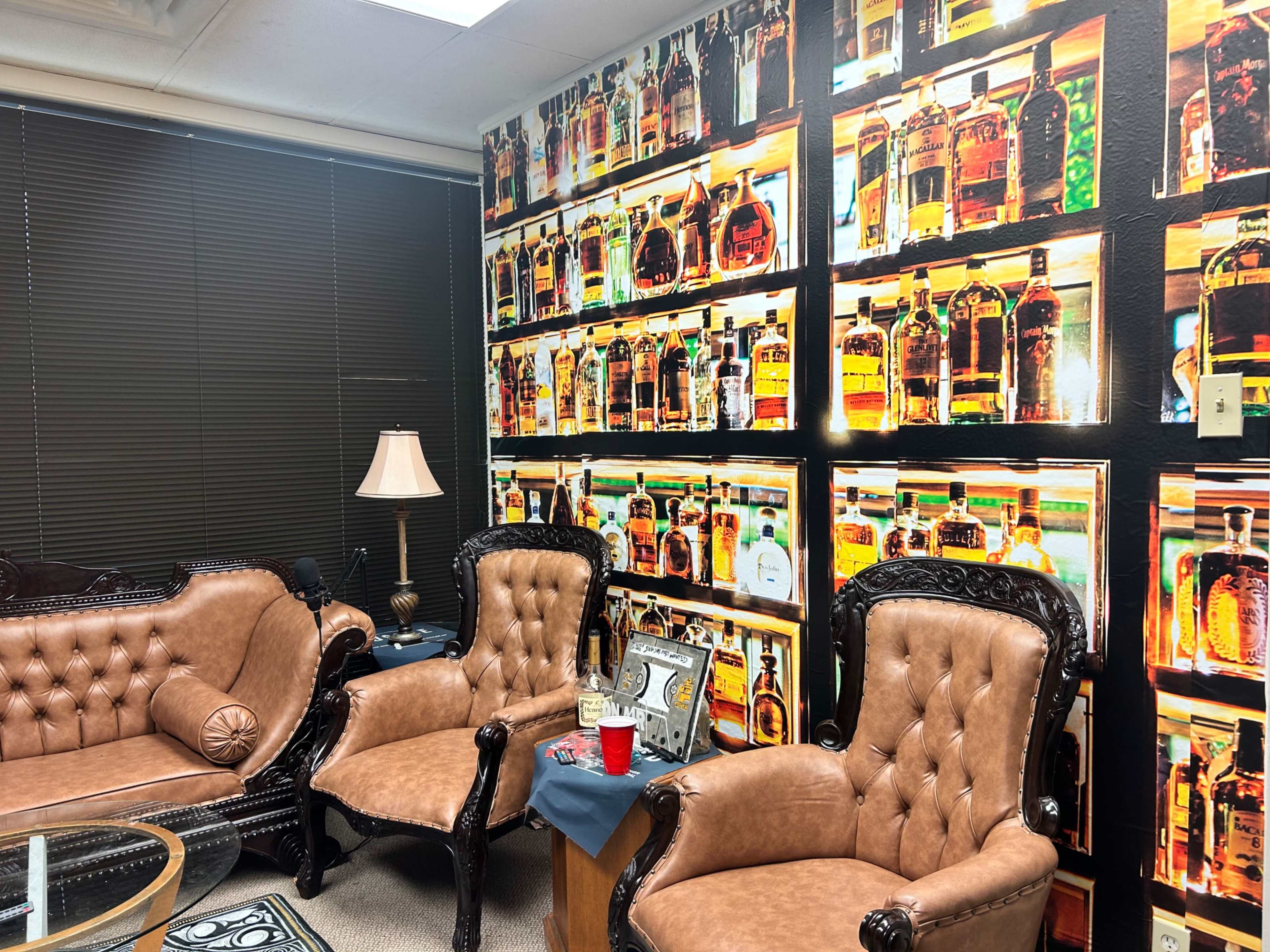 The room features two ornate brown leather chairs and a side table, with a wall covered in a mural of shelves filled with various bottles.