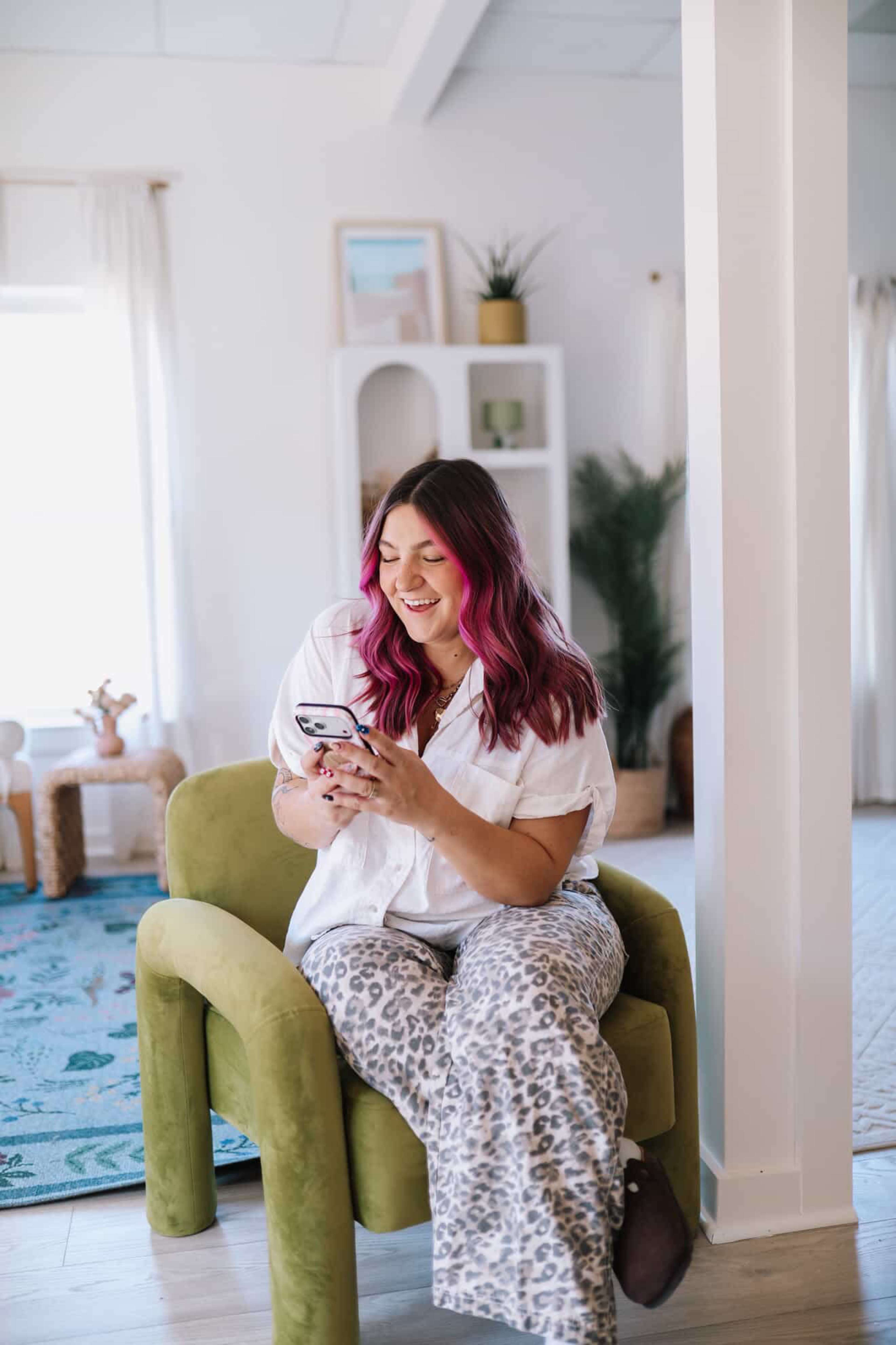 A woman with pink hair sits on a green chair, smiling while looking at her phone in a brightly lit room.