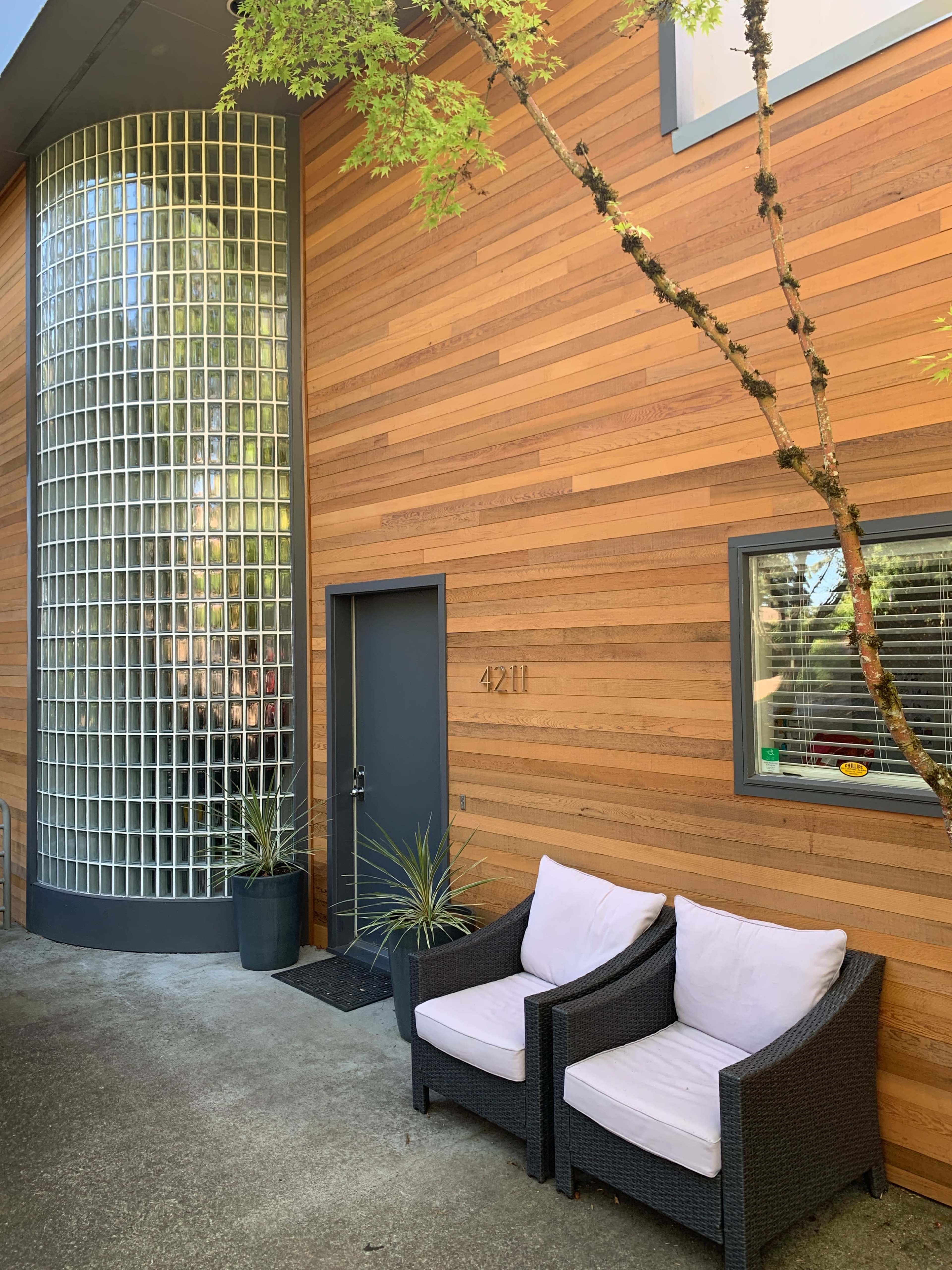 A modern building features a wooden exterior, large glass block wall, and two wicker chairs positioned near the entrance.