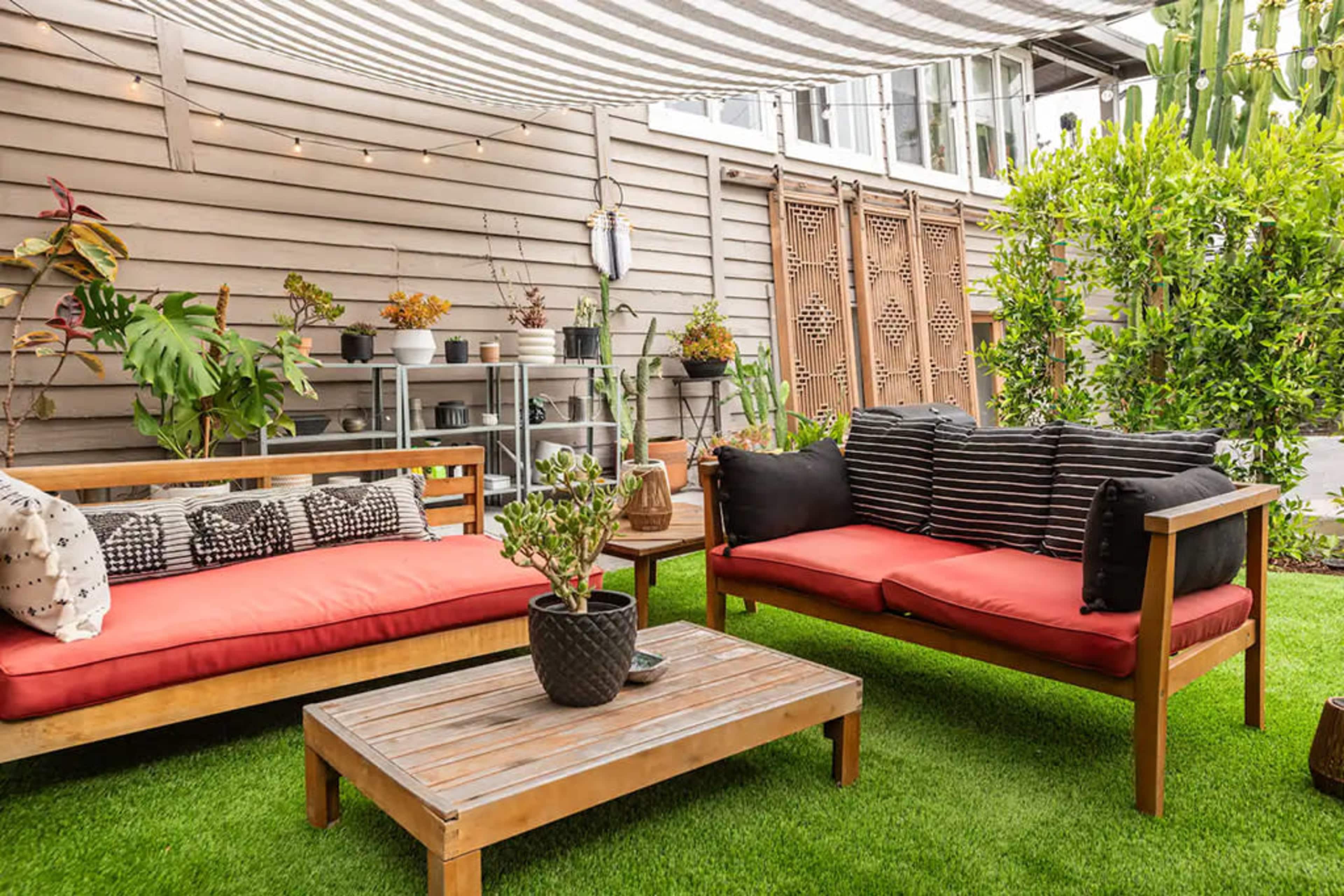 The image shows a cozy outdoor seating area with two wooden couches featuring red cushions, a wooden coffee table, and decorative plants under a striped canopy.