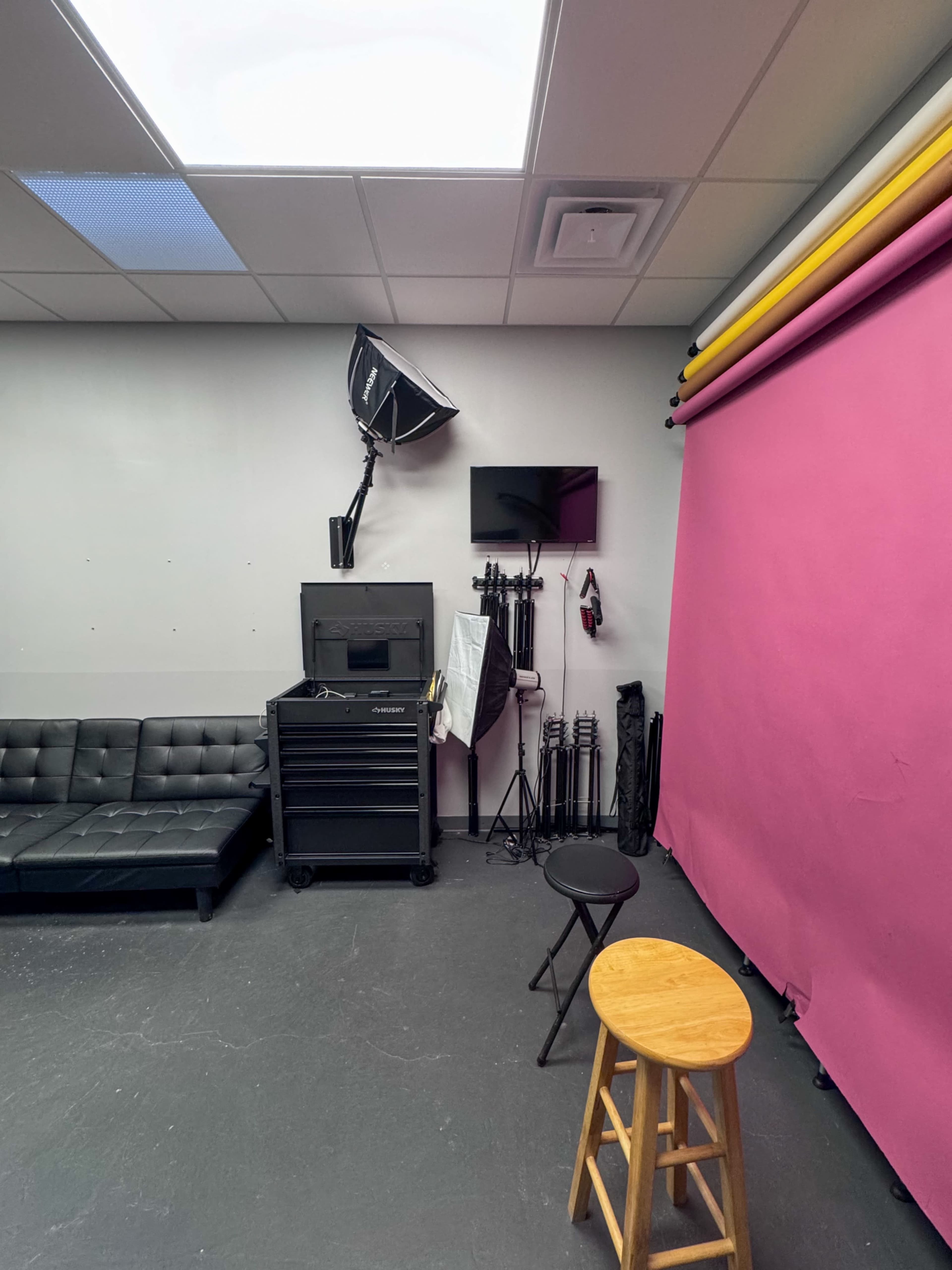 The image shows a photography studio with a pink backdrop, a couch, a wooden stool, and various lighting equipment.