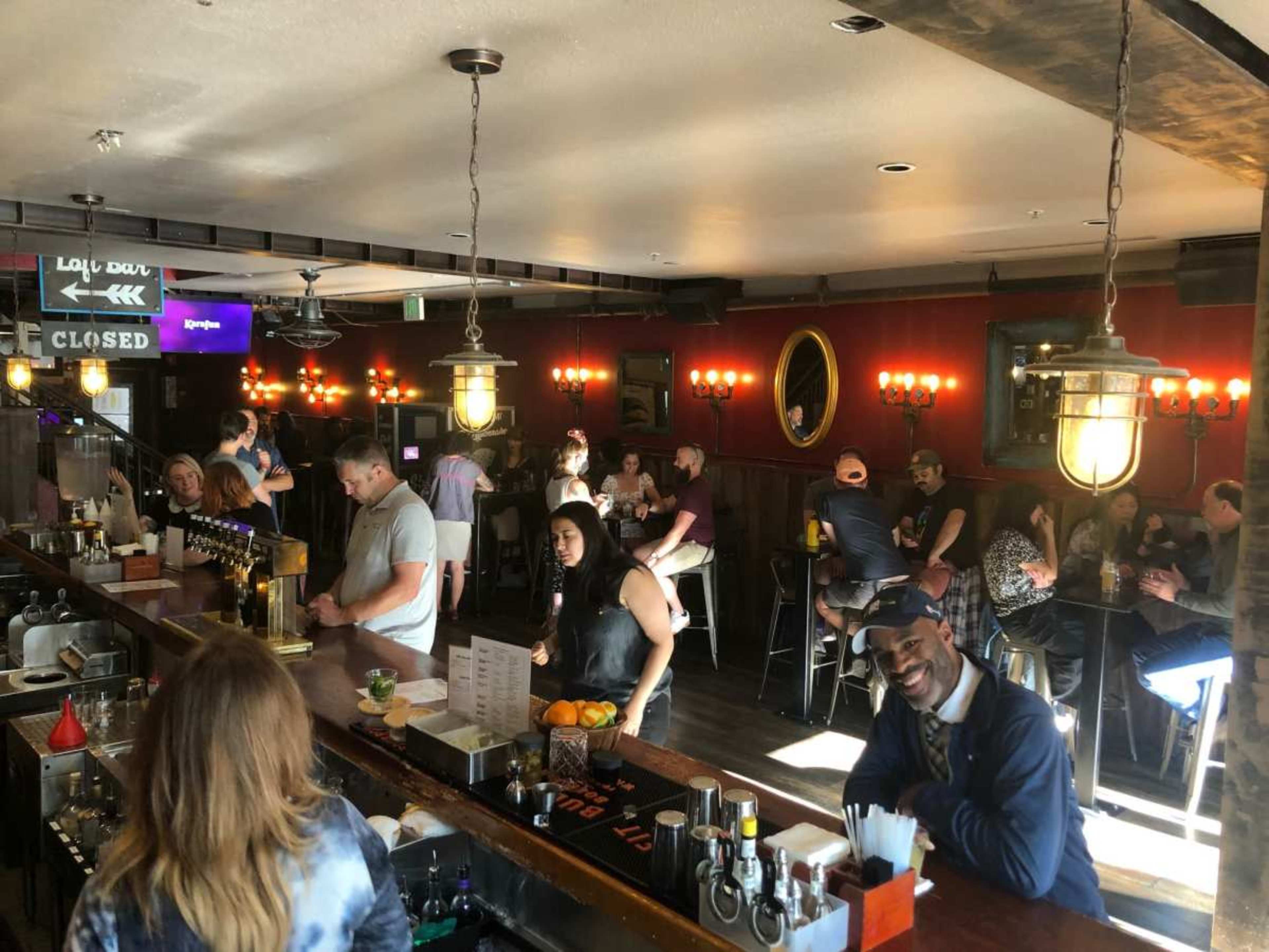 A bustling bar scene features patrons seated and socializing at tables and the bar, with a bartender preparing drinks in the foreground.