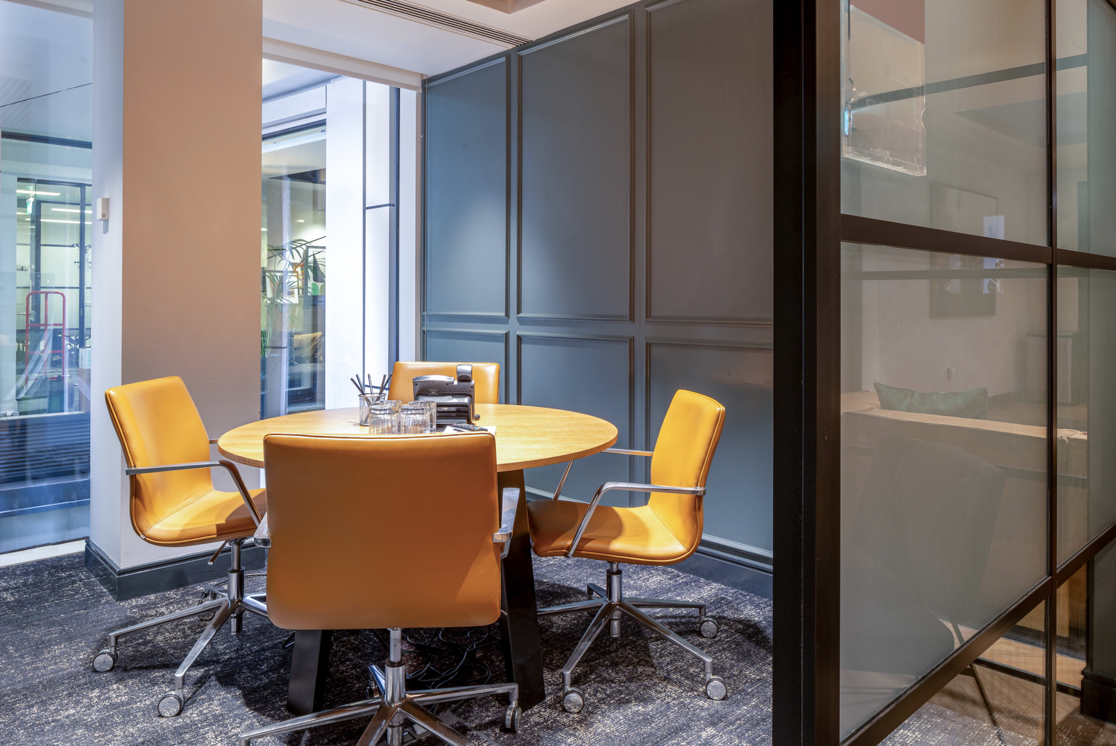 A small conference room features a round wooden table surrounded by four orange chairs, with large windows allowing natural light.