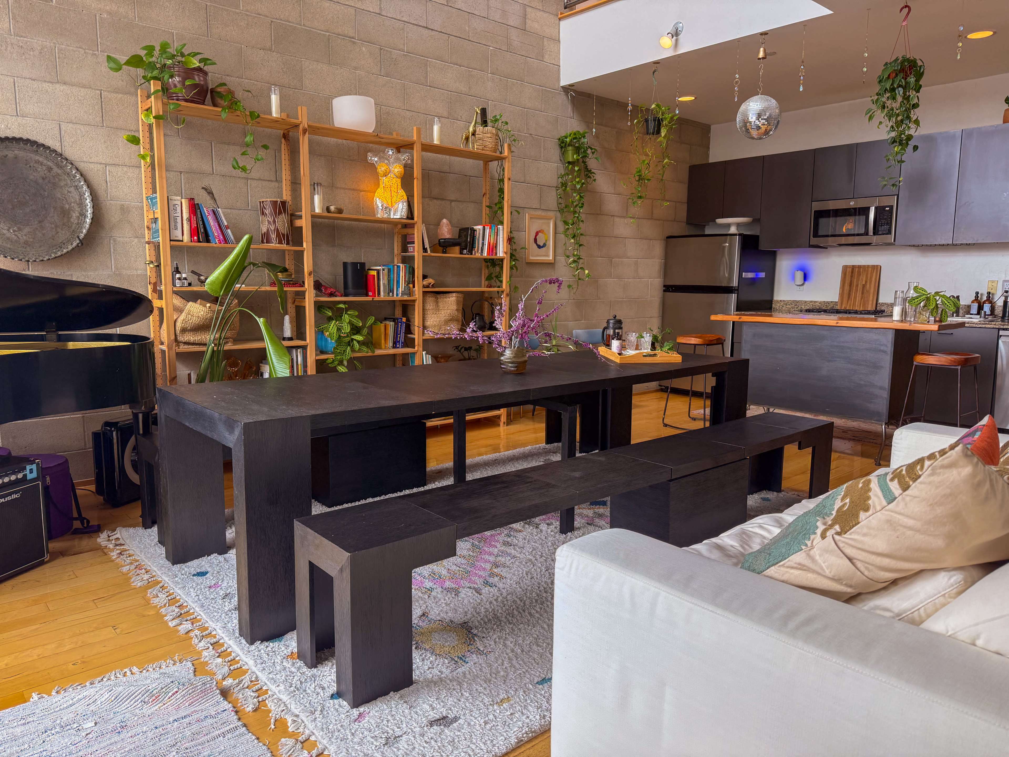The image shows a modern dining area with a large black table, matching benches, a wooden bookshelf filled with books and plants, and a kitchen in the background.