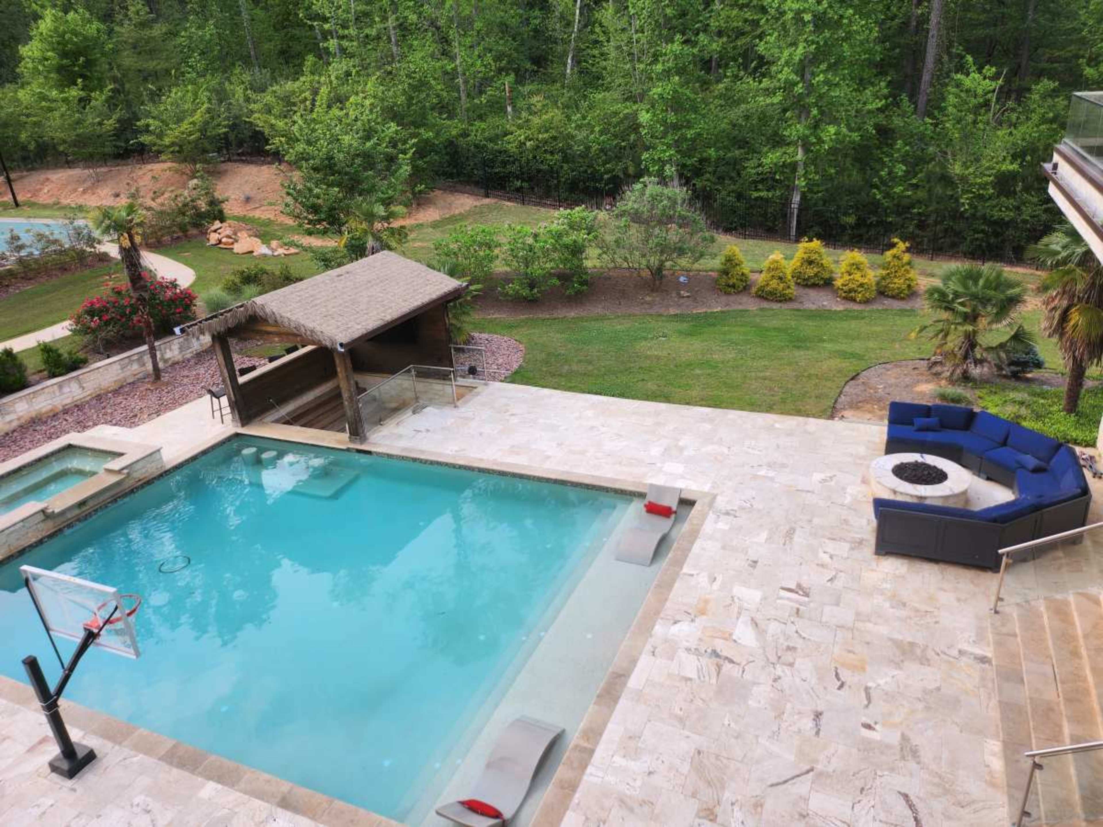 The image shows a backyard pool area with a blue pool, a seating area with a circular fire pit, and a basketball hoop near the water's edge, all surrounded by lush greenery.
