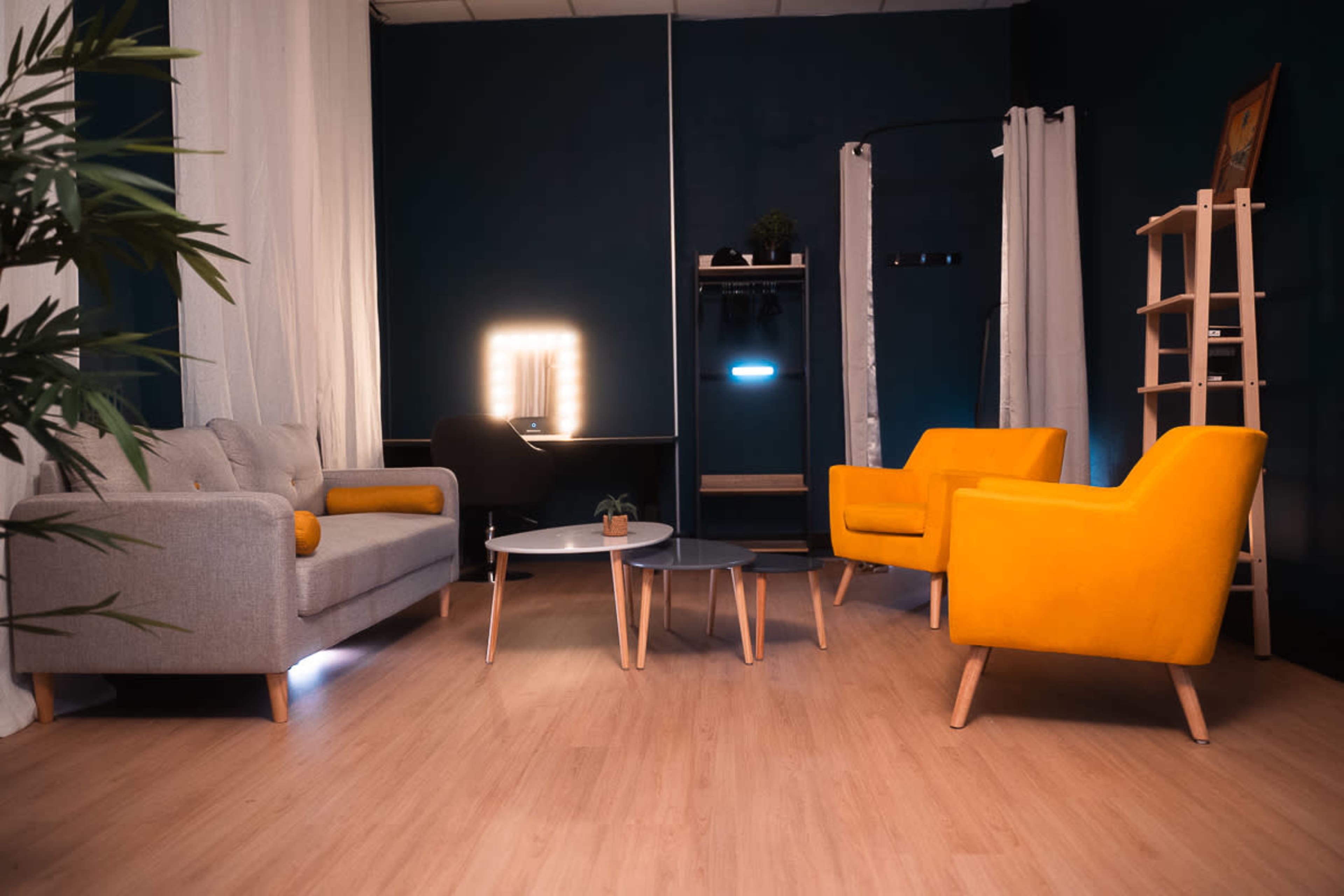 A modern interior space featuring a gray sofa, two yellow chairs, a round coffee table, and a mirror with lighting against a dark blue wall.
