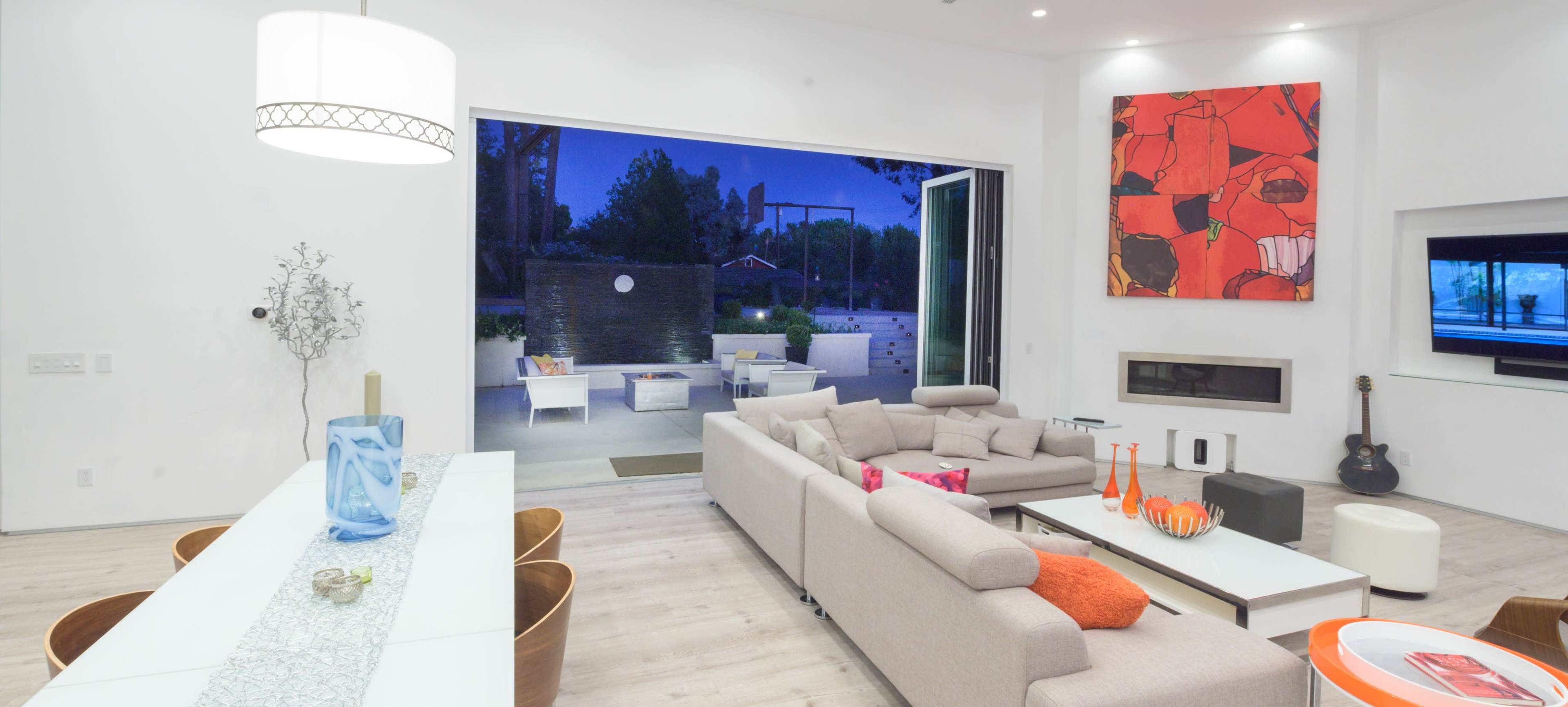 A modern living room with a light-colored sofa, a glass dining table, and large windows that open to an outdoor patio area.