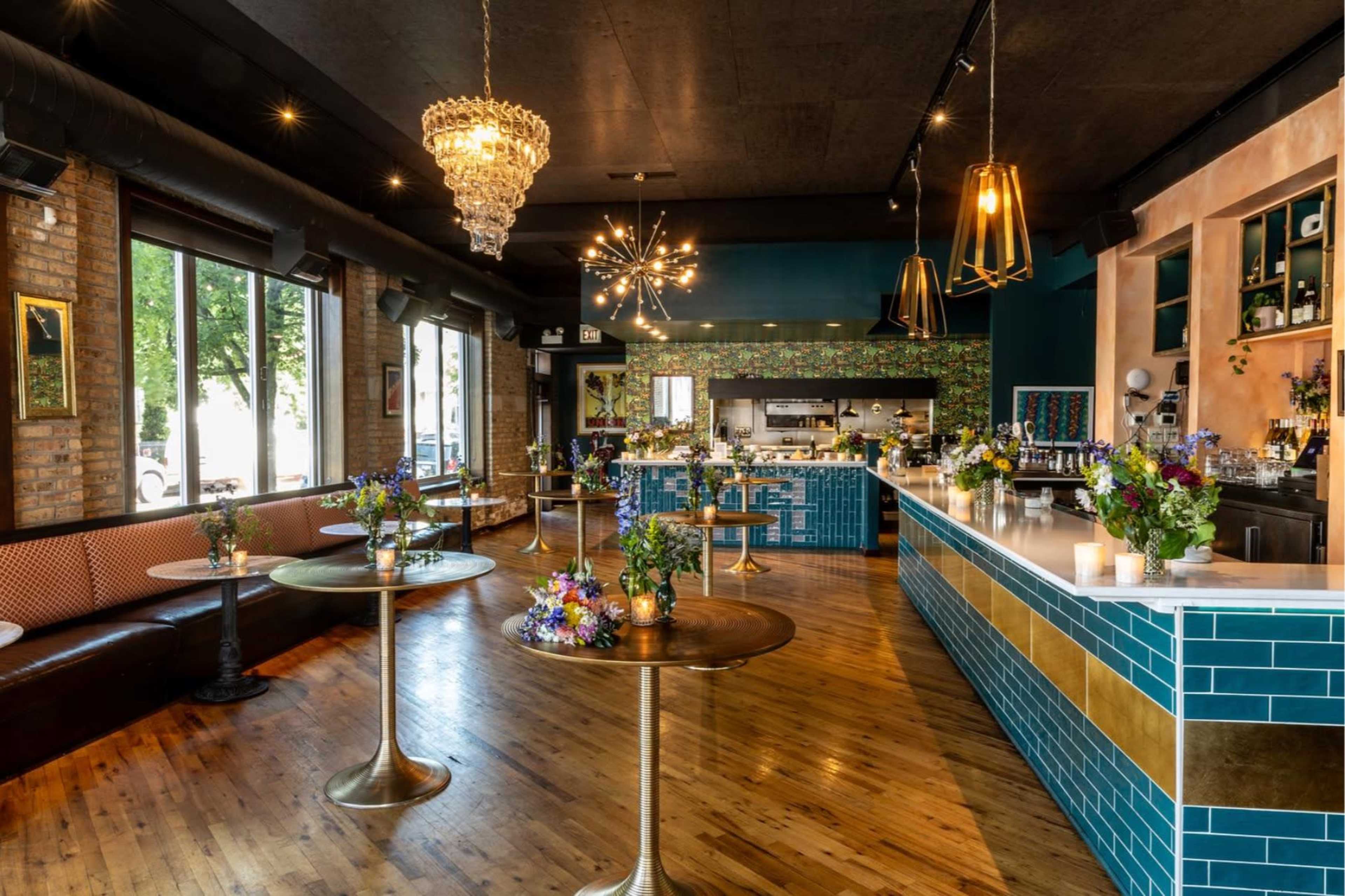 The image depicts a stylish restaurant interior featuring a bar area, wooden floors, colorful decor, and tables adorned with floral arrangements.