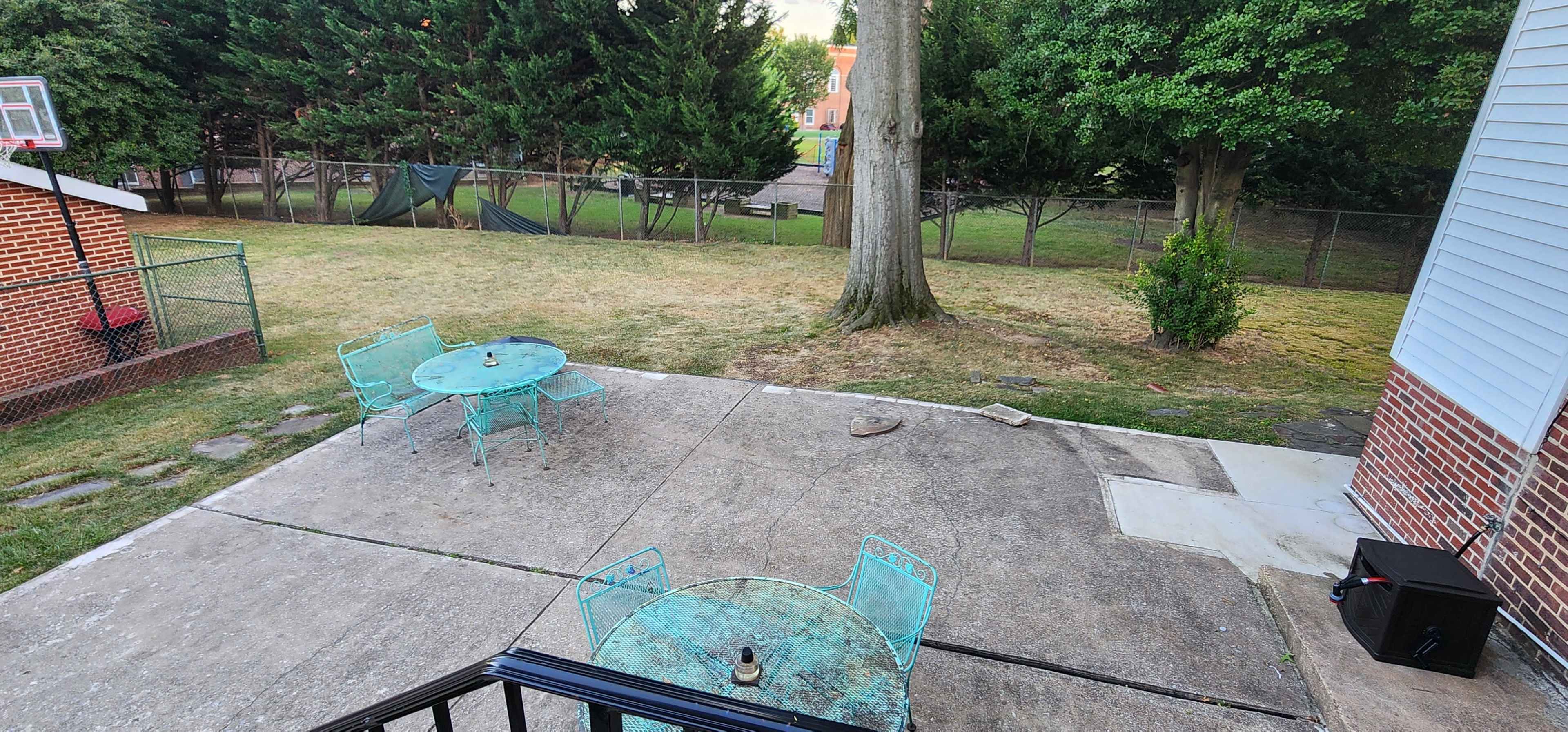 The image shows a backyard with two metal tables and chairs on a concrete patio, surrounded by a grassy area and trees.