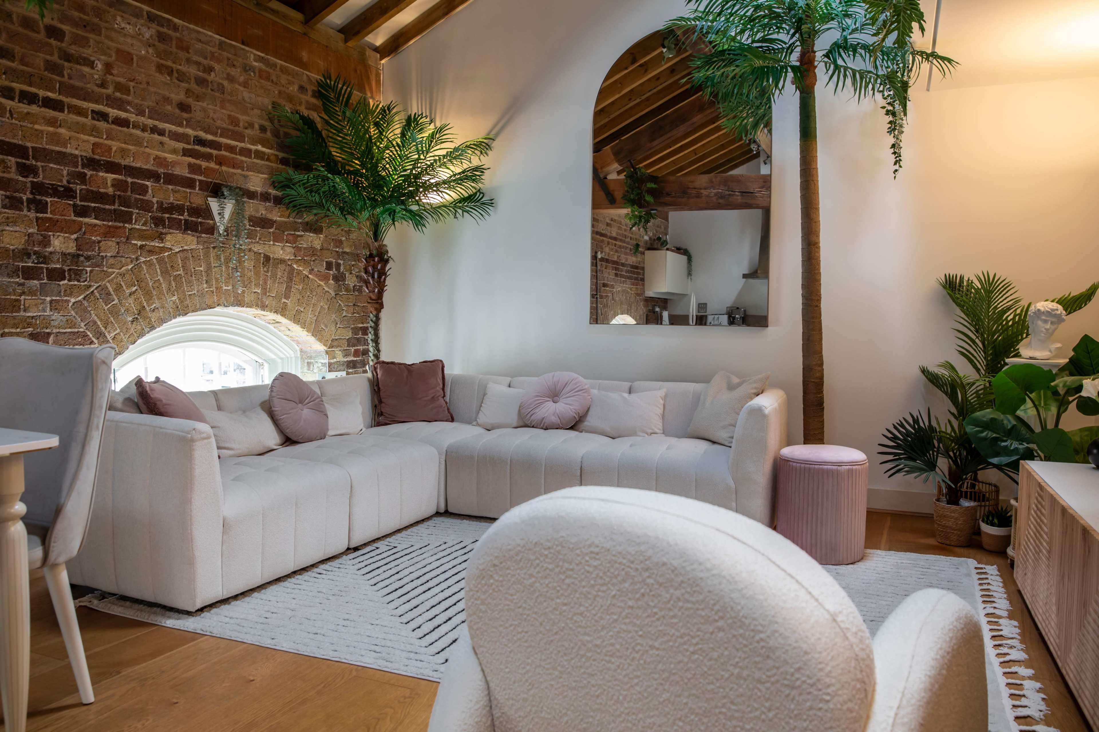 A spacious living area features a curved white sofa, a wooden ceiling, and decorative plants beside a large arched window.
