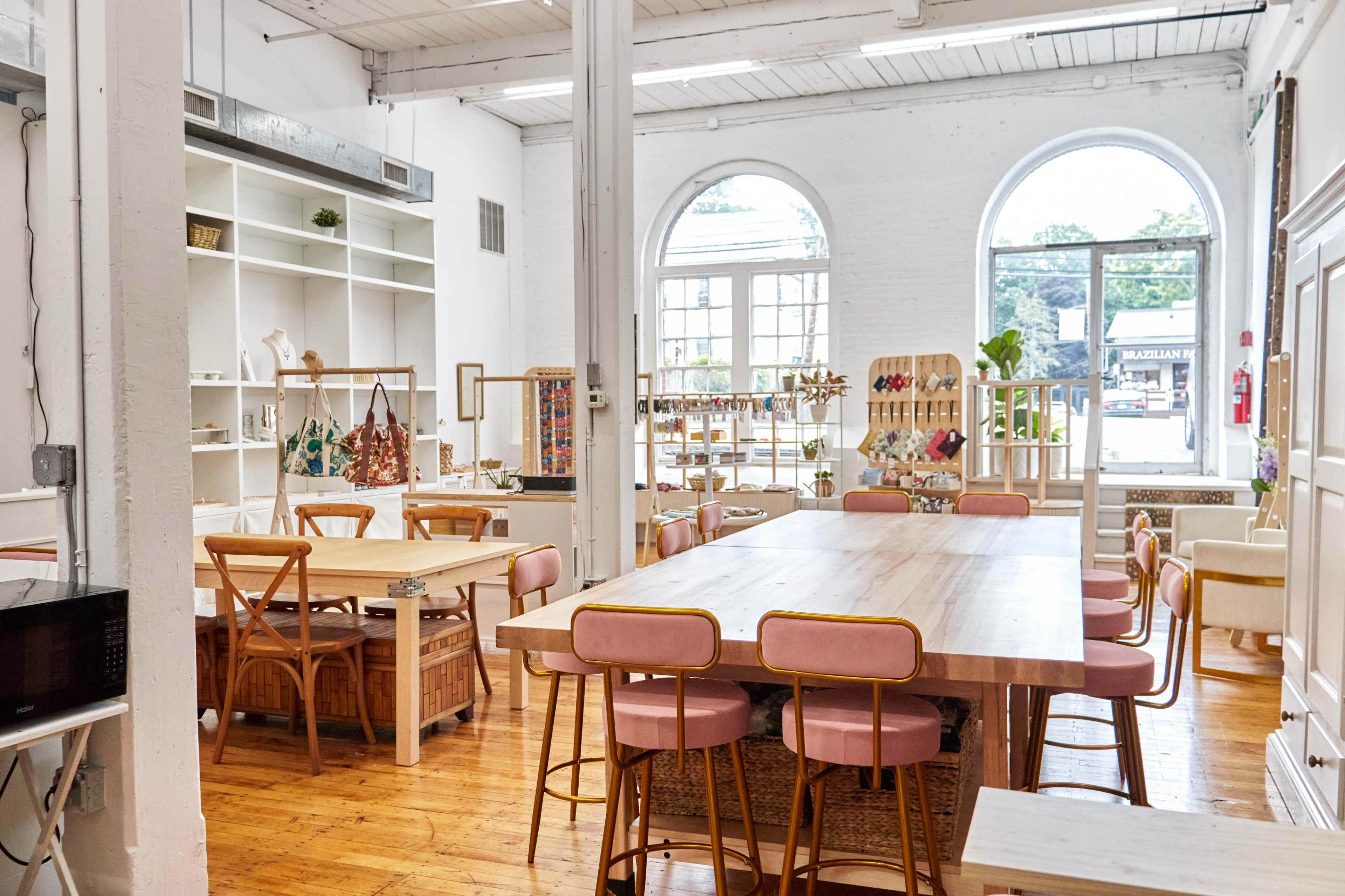 A bright, open workspace with wooden tables, pink chairs, and shelves displaying various decorative items.