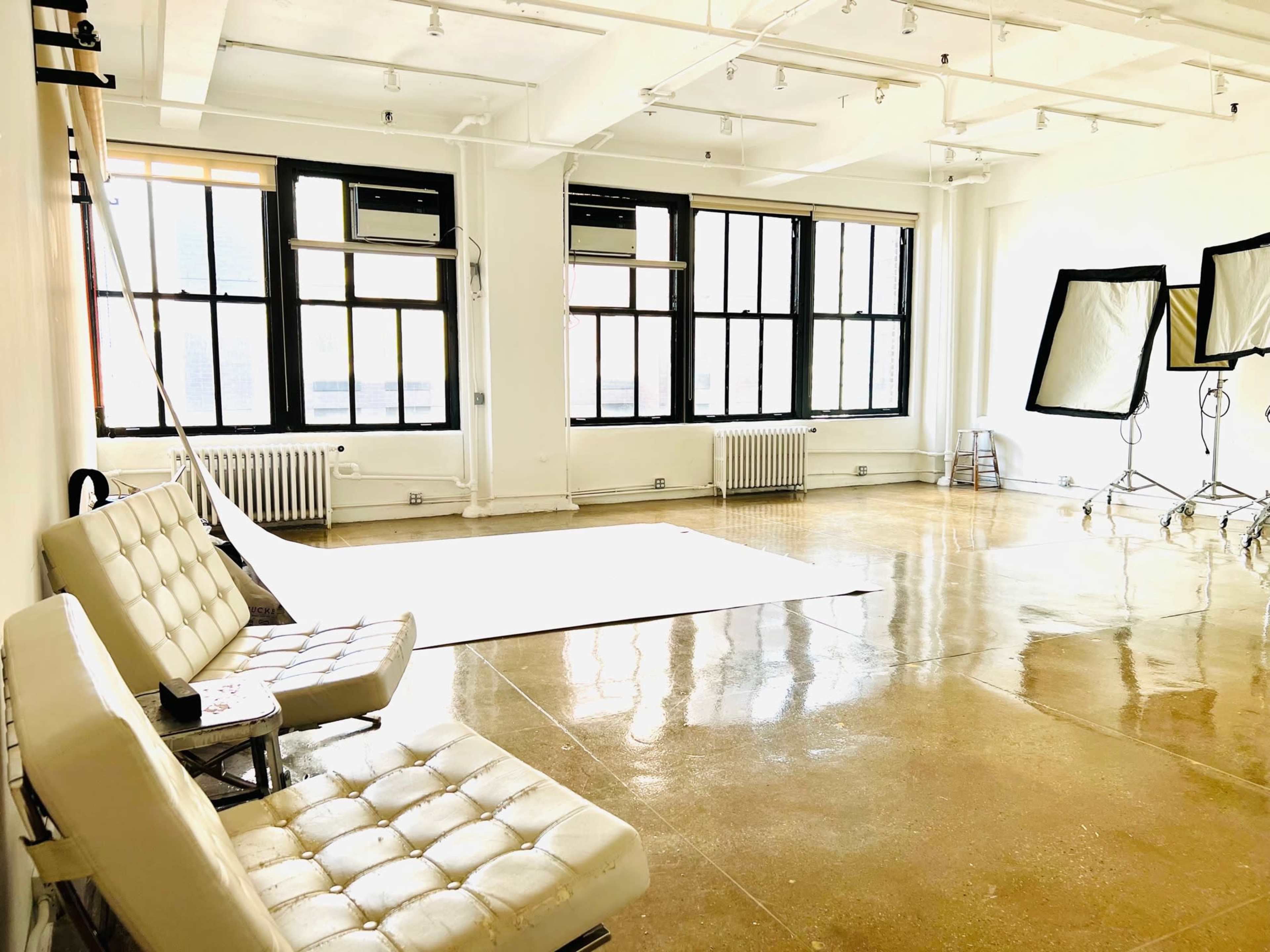 The image shows a bright photography studio with large windows, two white couches, and equipment set up for a shoot.