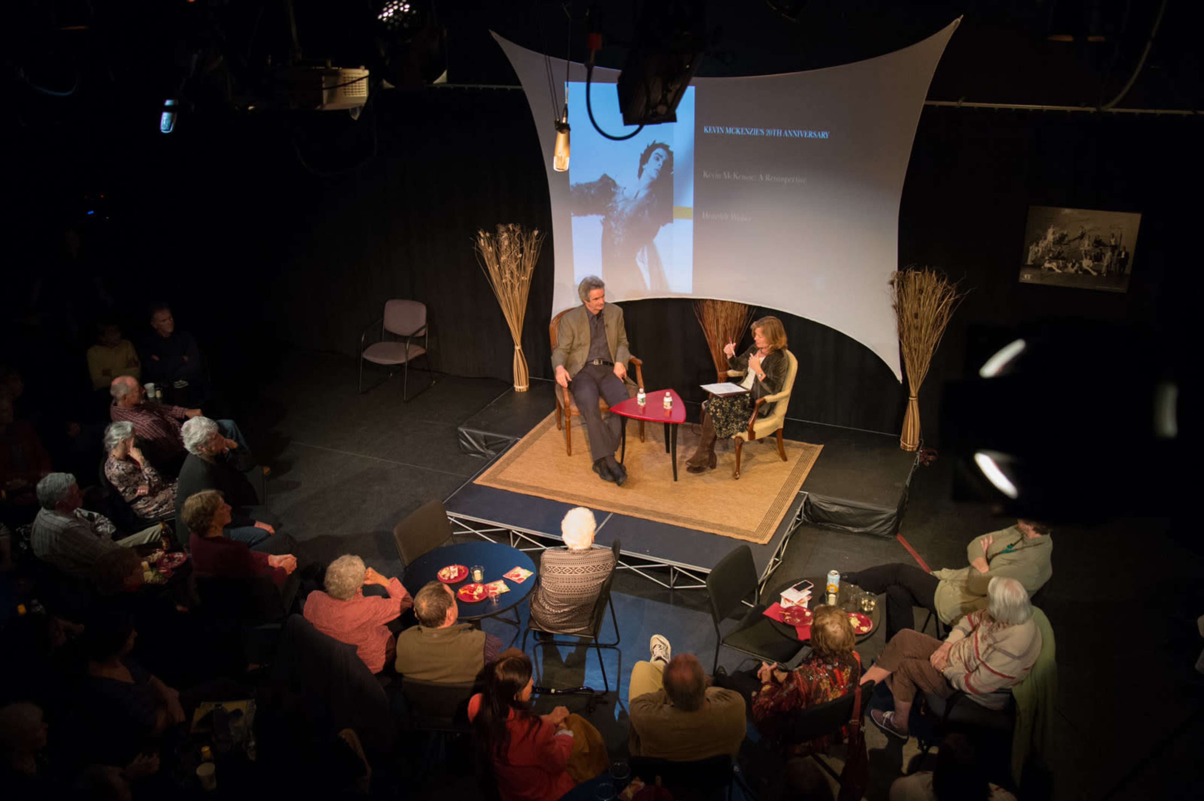 A panel discussion is taking place on a stage with a seated audience in a dimly lit room.