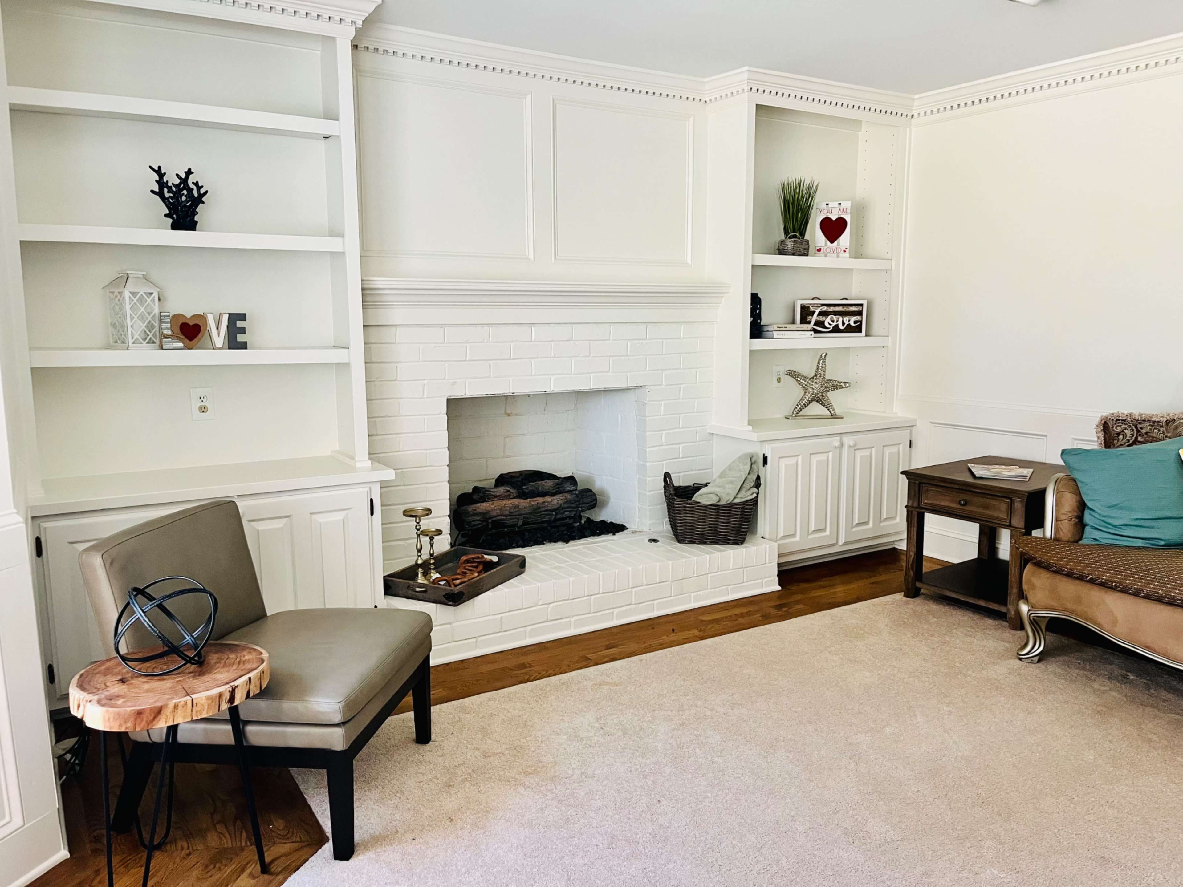 A cozy, well-decorated living room featuring a white brick fireplace, built-in shelves with decorative items, a beige rug, and a seating area with a wooden side table and a chair.