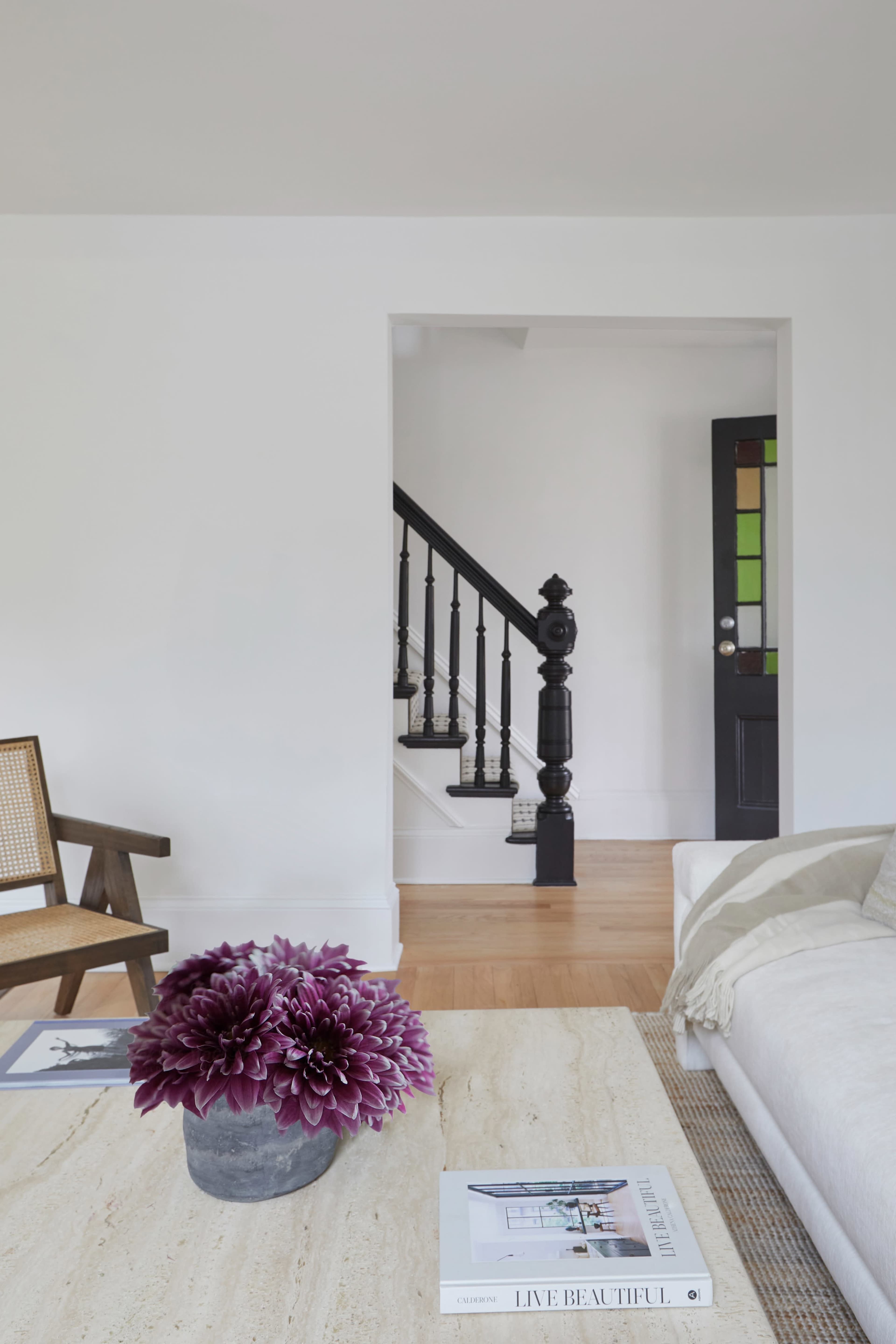 A minimalist living room features a large flower arrangement on a table, with a staircase visible in the background.
