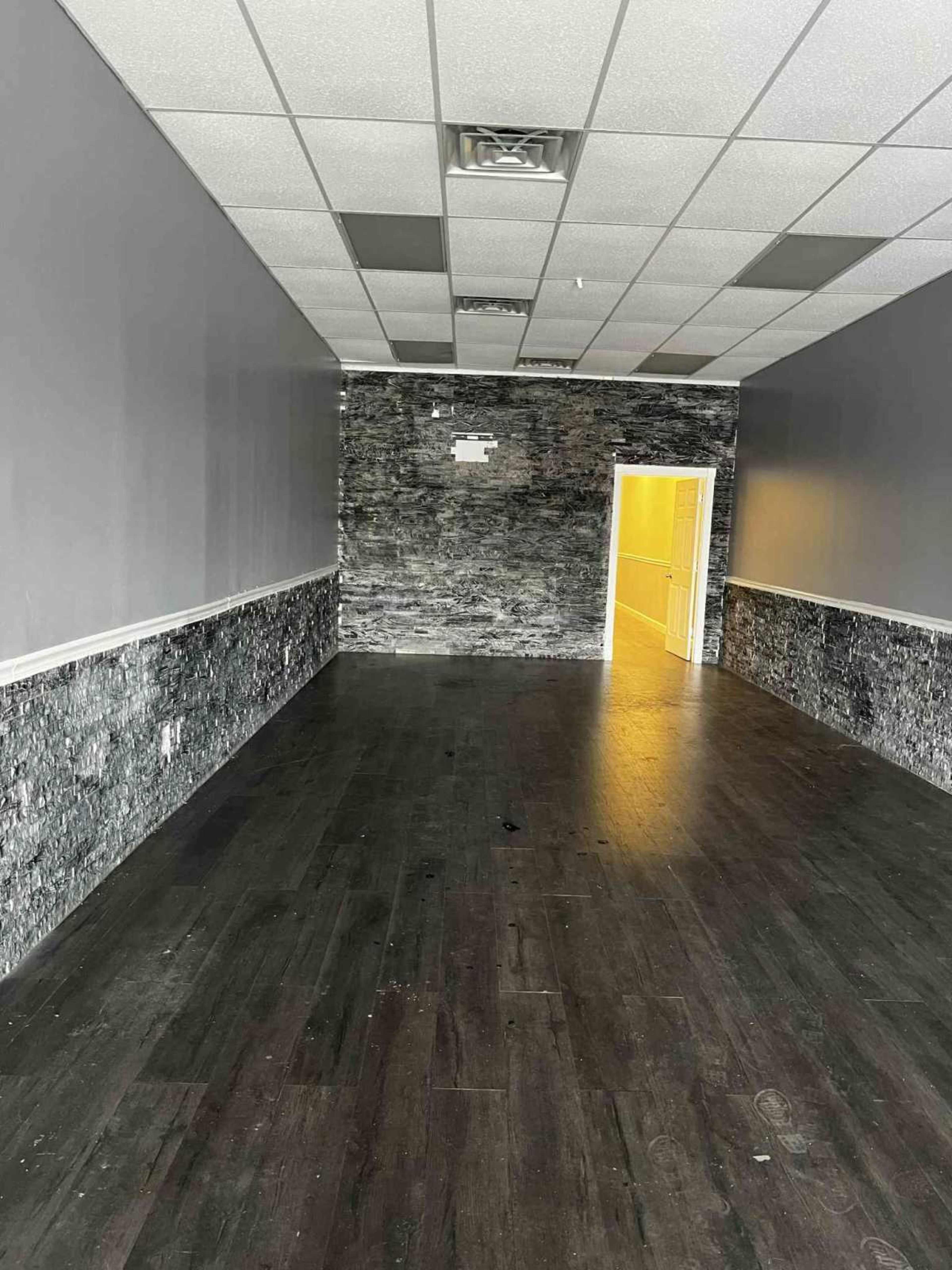 The image shows an empty room with dark wooden flooring and gray walls featuring a textured pattern, leading to a doorway with warm yellow light at the end of a narrow hallway.