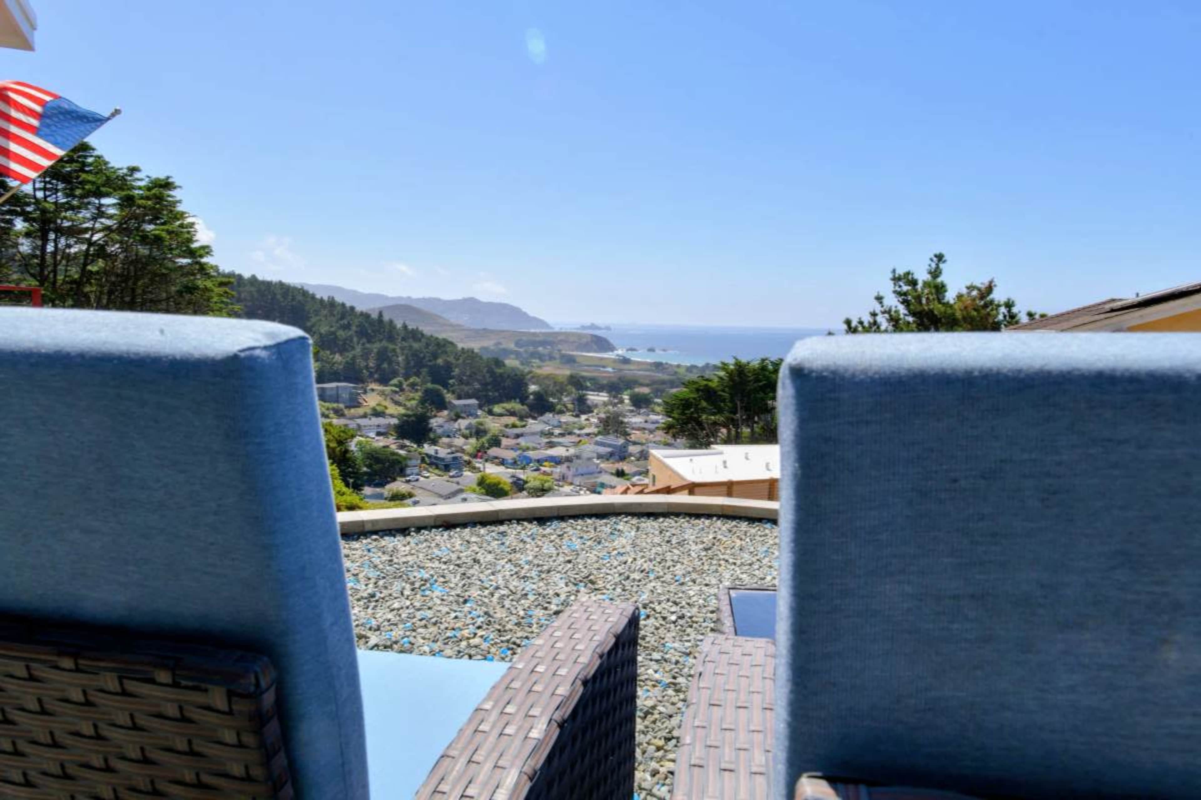 The image shows two blue chairs positioned on a terrace overlooking a coastal view with hills and a town in the distance.