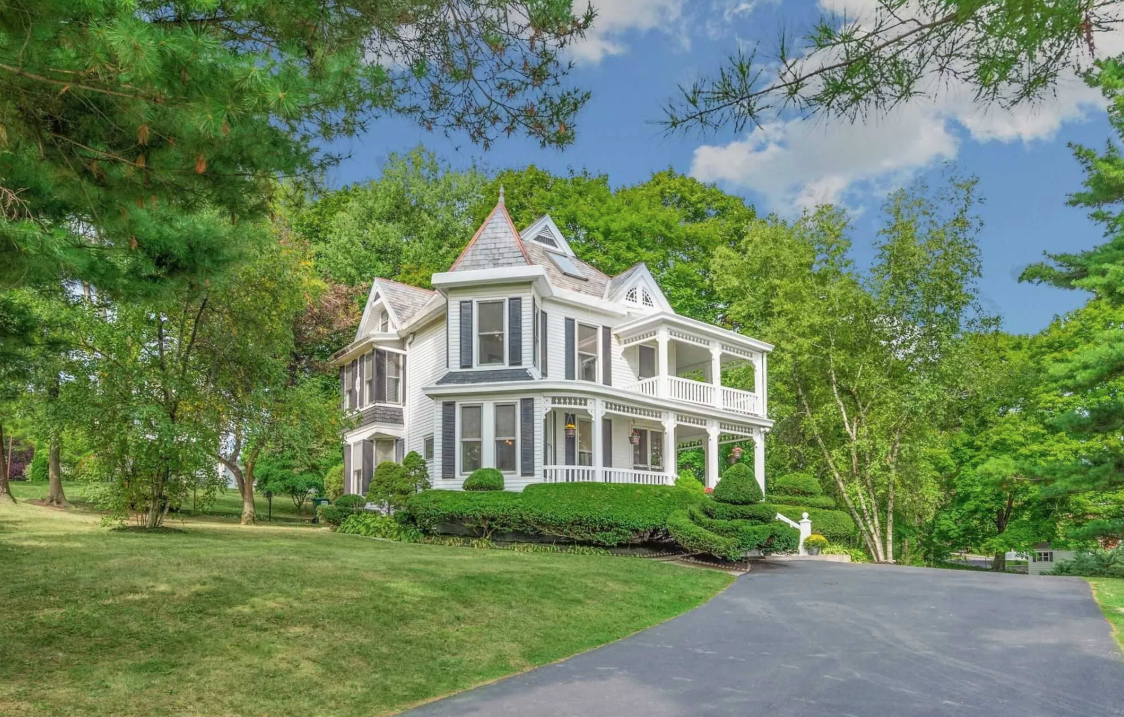 The image shows a two-story Victorian-style house with a wraparound porch, surrounded by lush green trees and lawn.