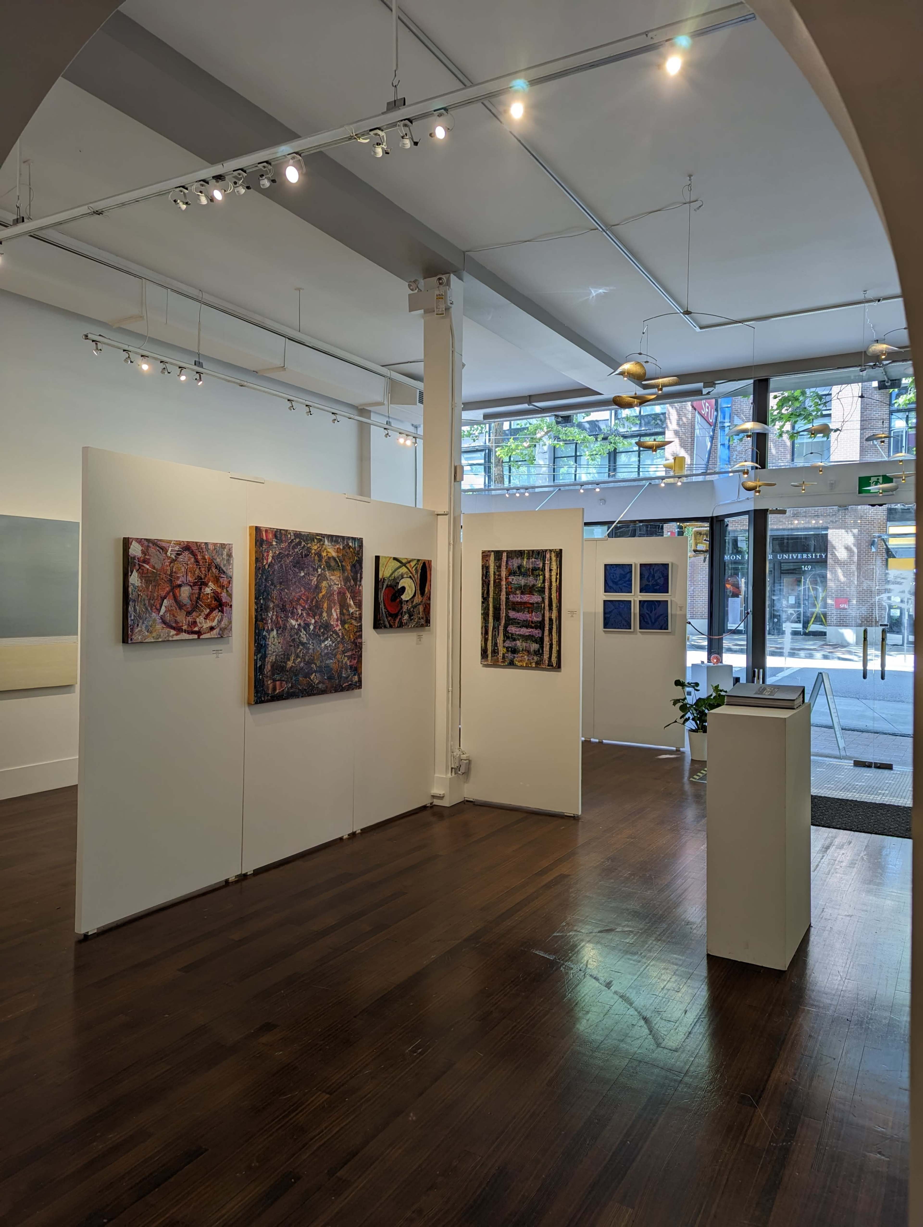 An art gallery with white walls displaying colorful paintings, while large windows allow natural light to flood the space.